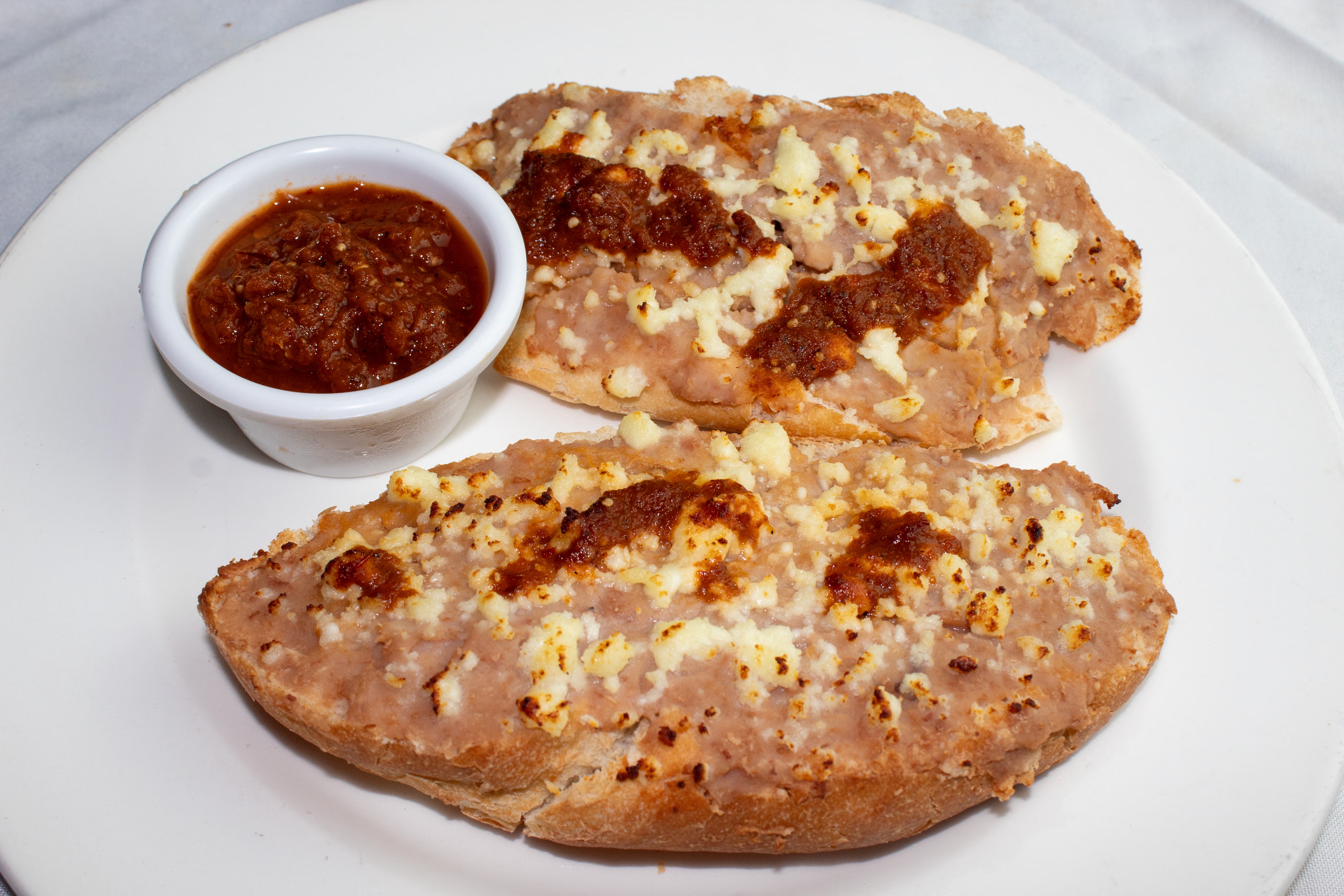 Two bread slices topped with refried beans, melted cheese, and salsa on a white plate, accompanied by a small bowl of salsa