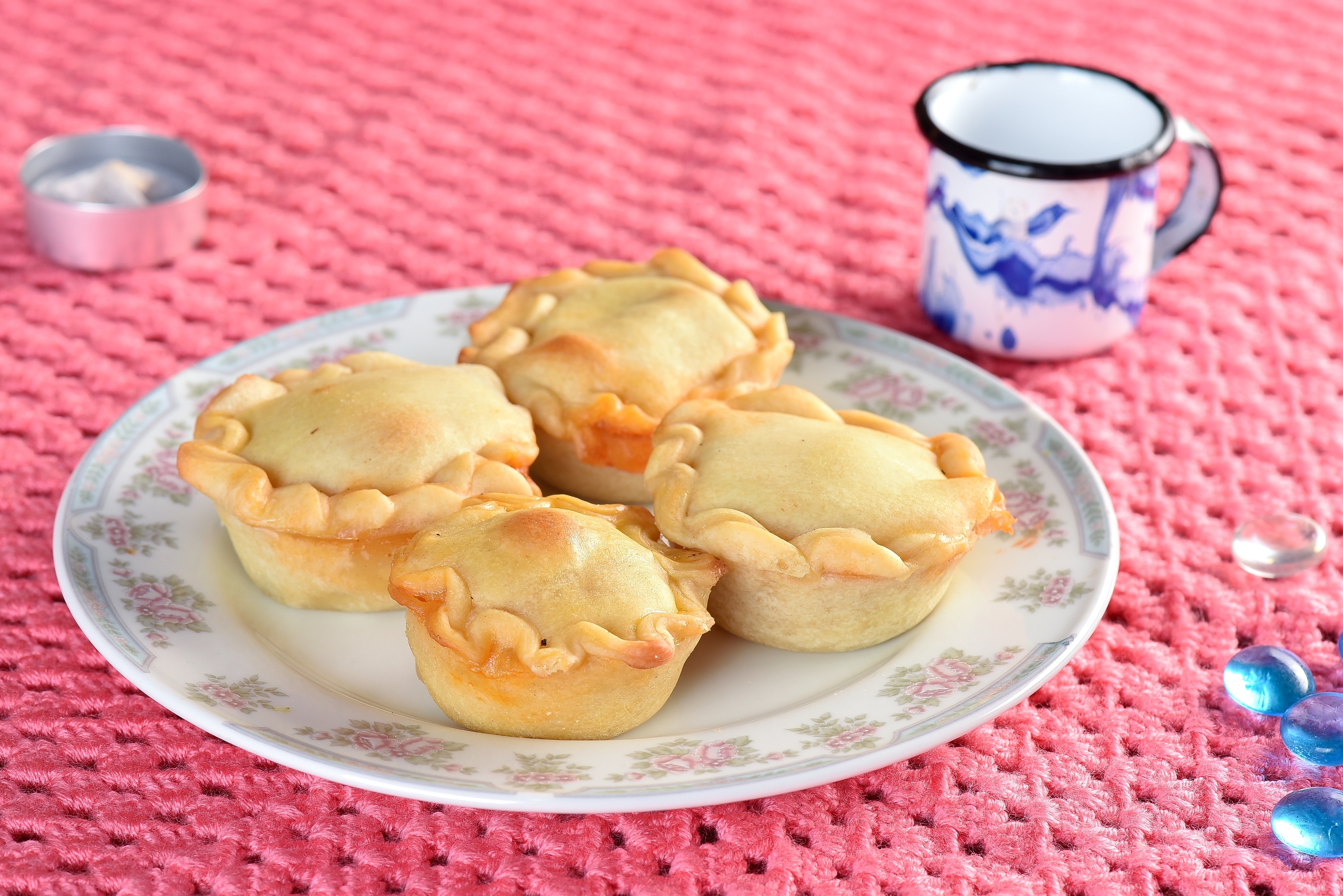 Four small, homemade pies on a floral plate next to a decorative mug and scattered gems on a textured surface