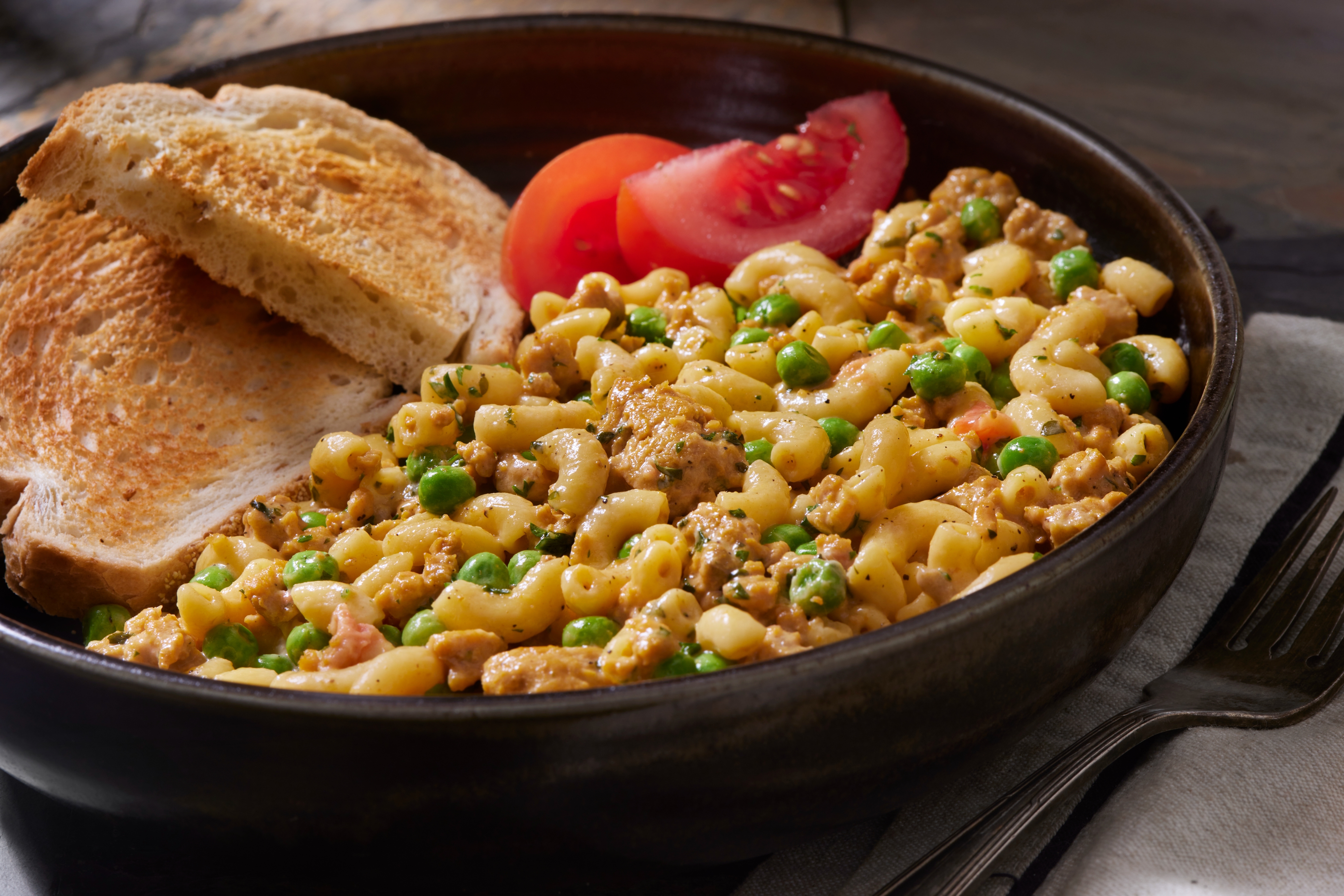 A bowl of macaroni mixed with peas and ground beef, served with sliced tomatoes and toasted bread on the side