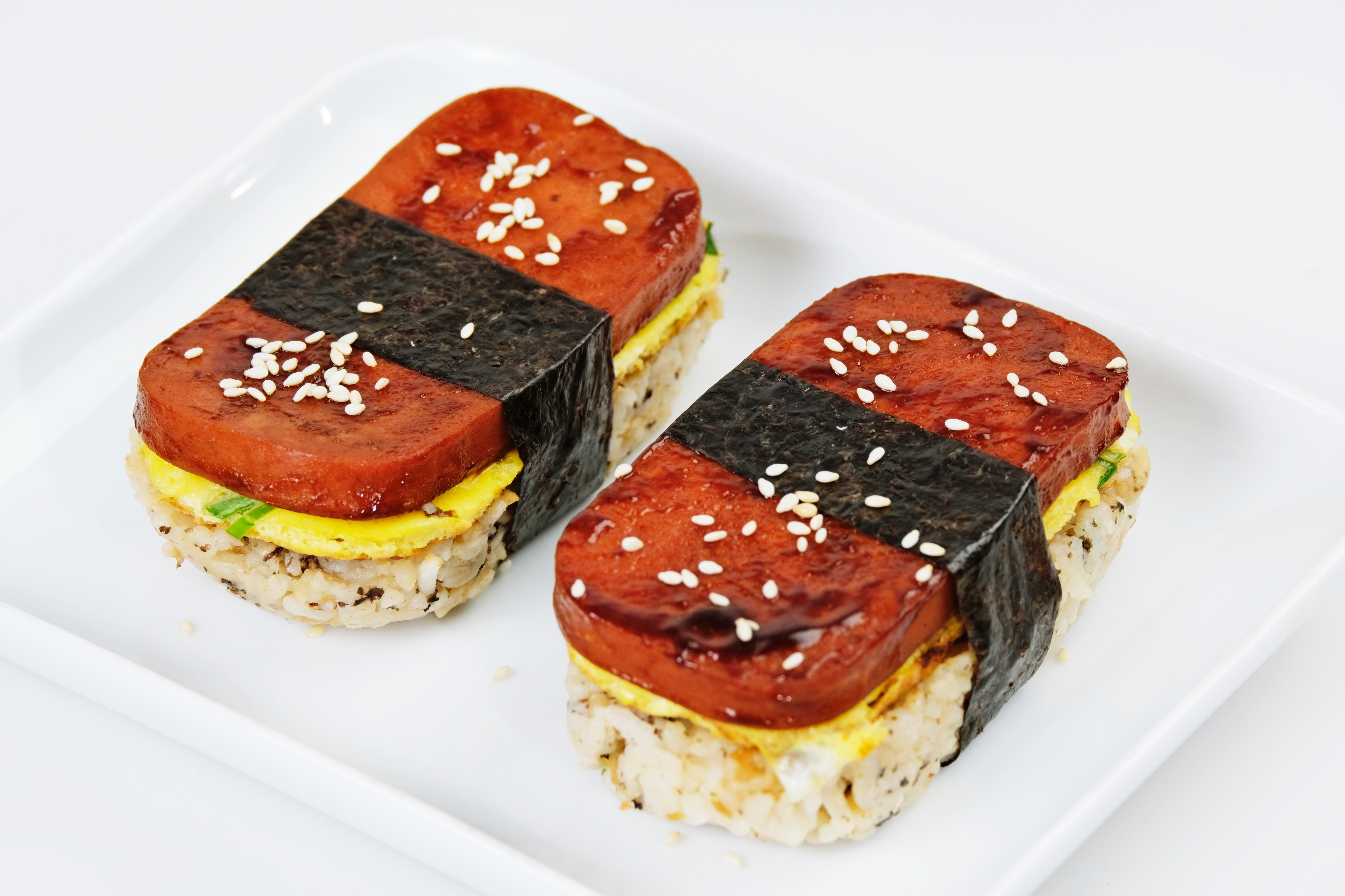 Two pieces of Spam musubi with rice, egg, and seaweed, topped with sesame seeds on a white plate