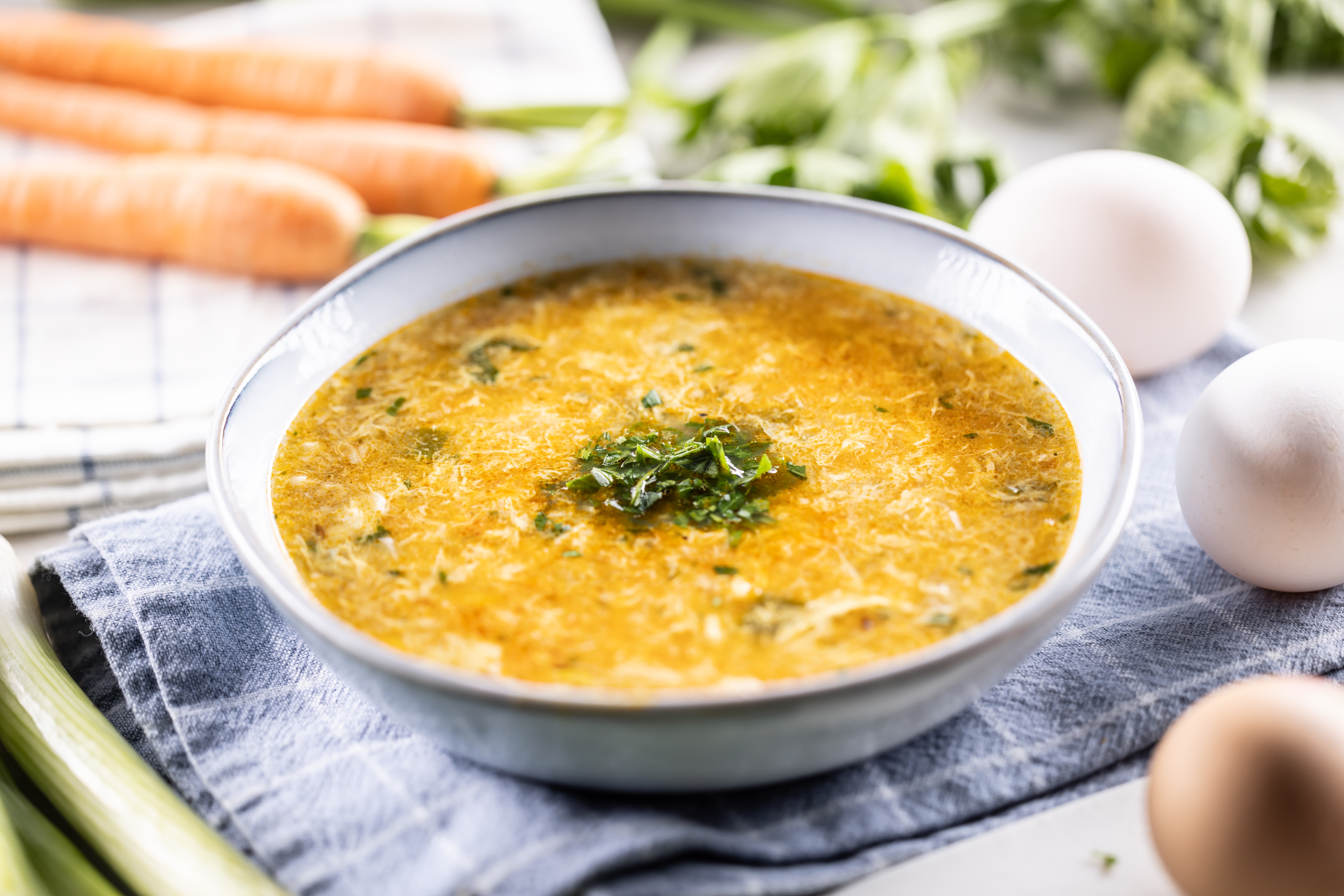 A bowl of egg drop soup garnished with herbs, surrounded by fresh vegetables and eggs on a cloth