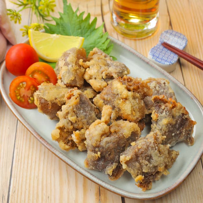 皿に盛られたこんにゃくの唐揚げ、トマト、レモン、青しそが置かれている。背景に飲み物。