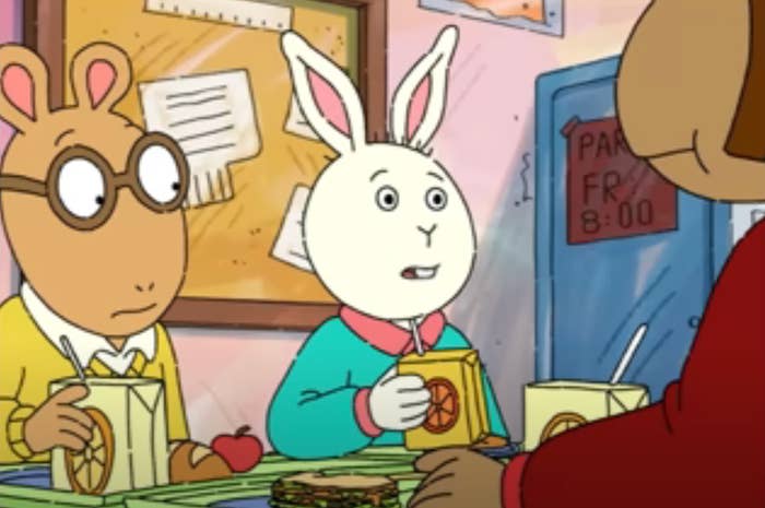 Animated characters at a school lunch table with juice boxes, a sandwich, and an apple, appearing engaged in conversation