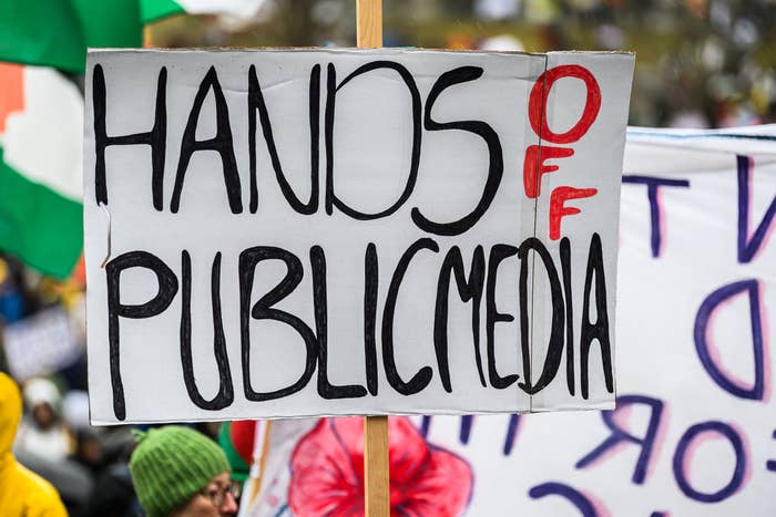 Sign reading &quot;Hands Off Public Media&quot; held at a protest, with people and other signs blurred in the background