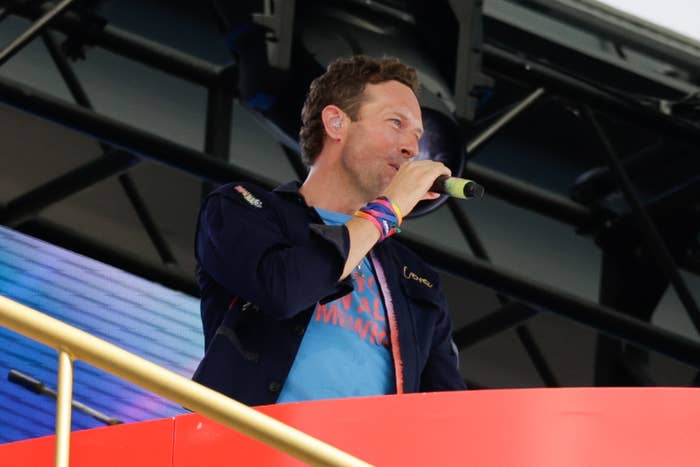 Chris Martin in a casual jacket and T-shirt, speaking into a microphone on an elevated platform