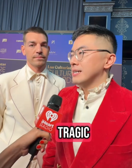 Two individuals on a press event stage. The person in front wears a red suit with a ruffled shirt, speaking into an iHeartRadio microphone. The word &quot;TRAGIC&quot; appears