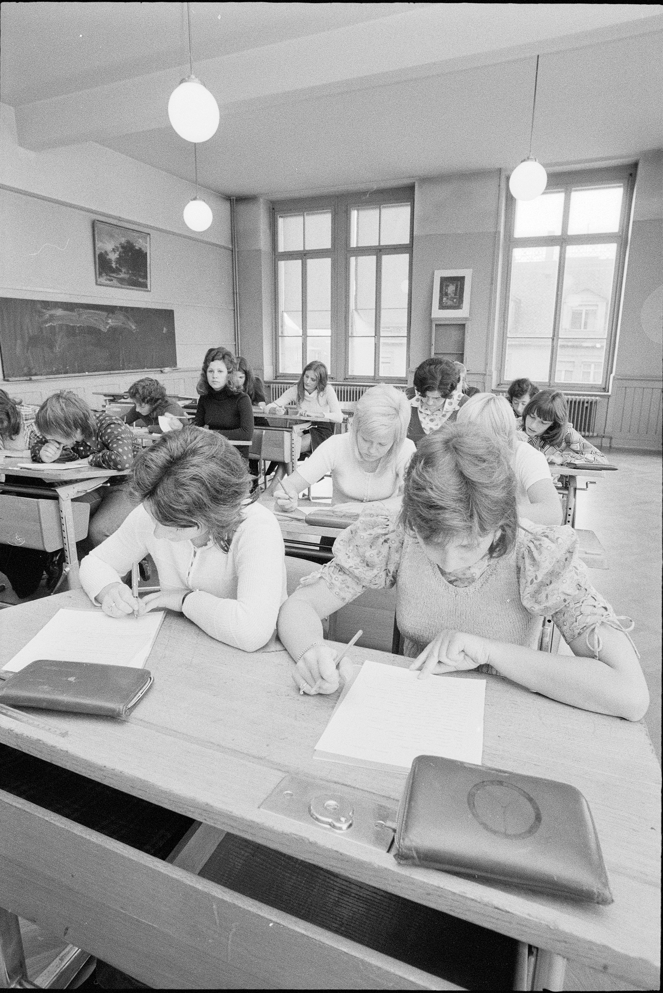 Students diligently writing at desks in a classroom with large windows and overhead lights