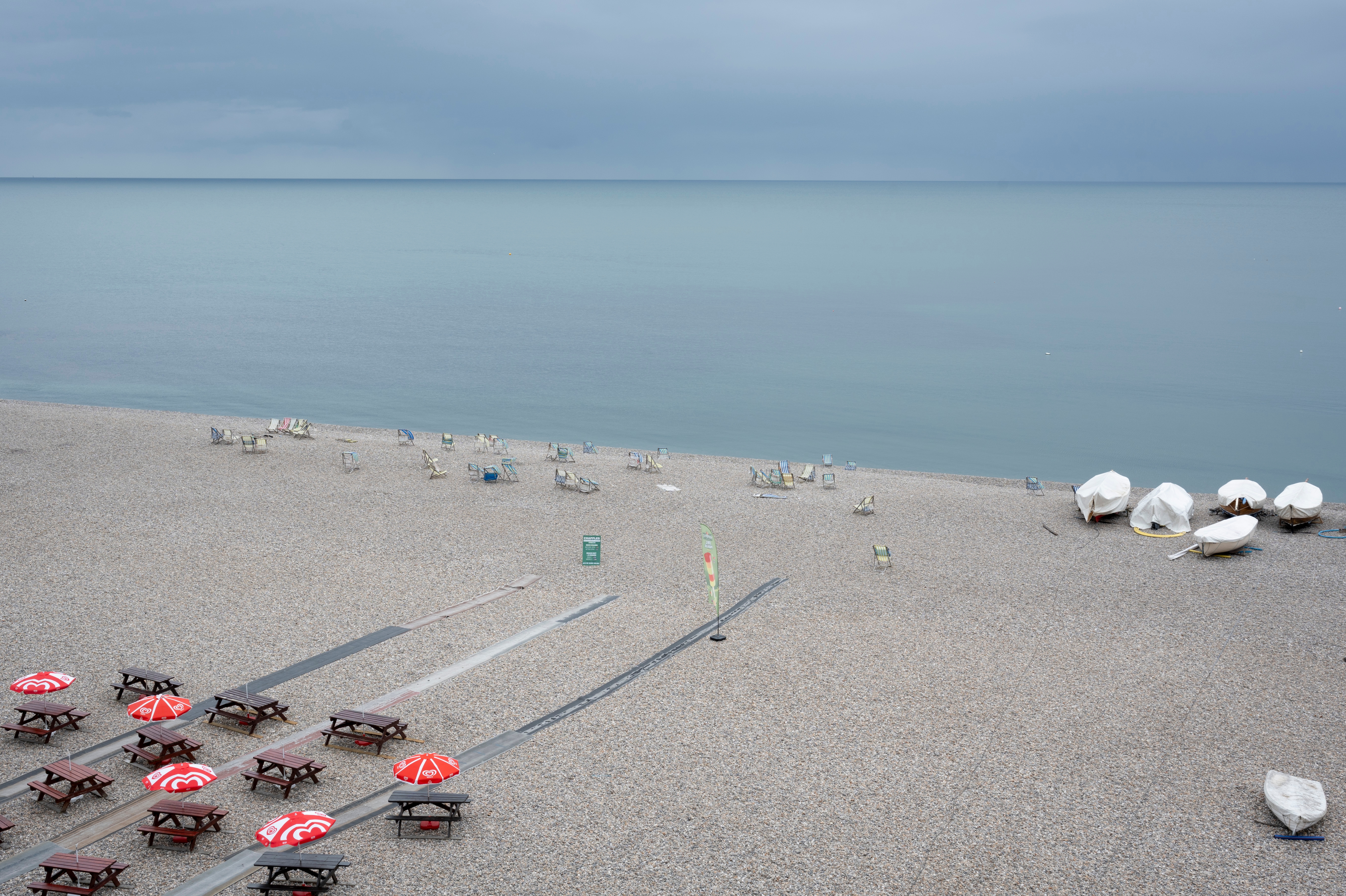 Empty pebble beach with rows of closed red umbrellas and sunbeds, near calm sea under a cloudy sky