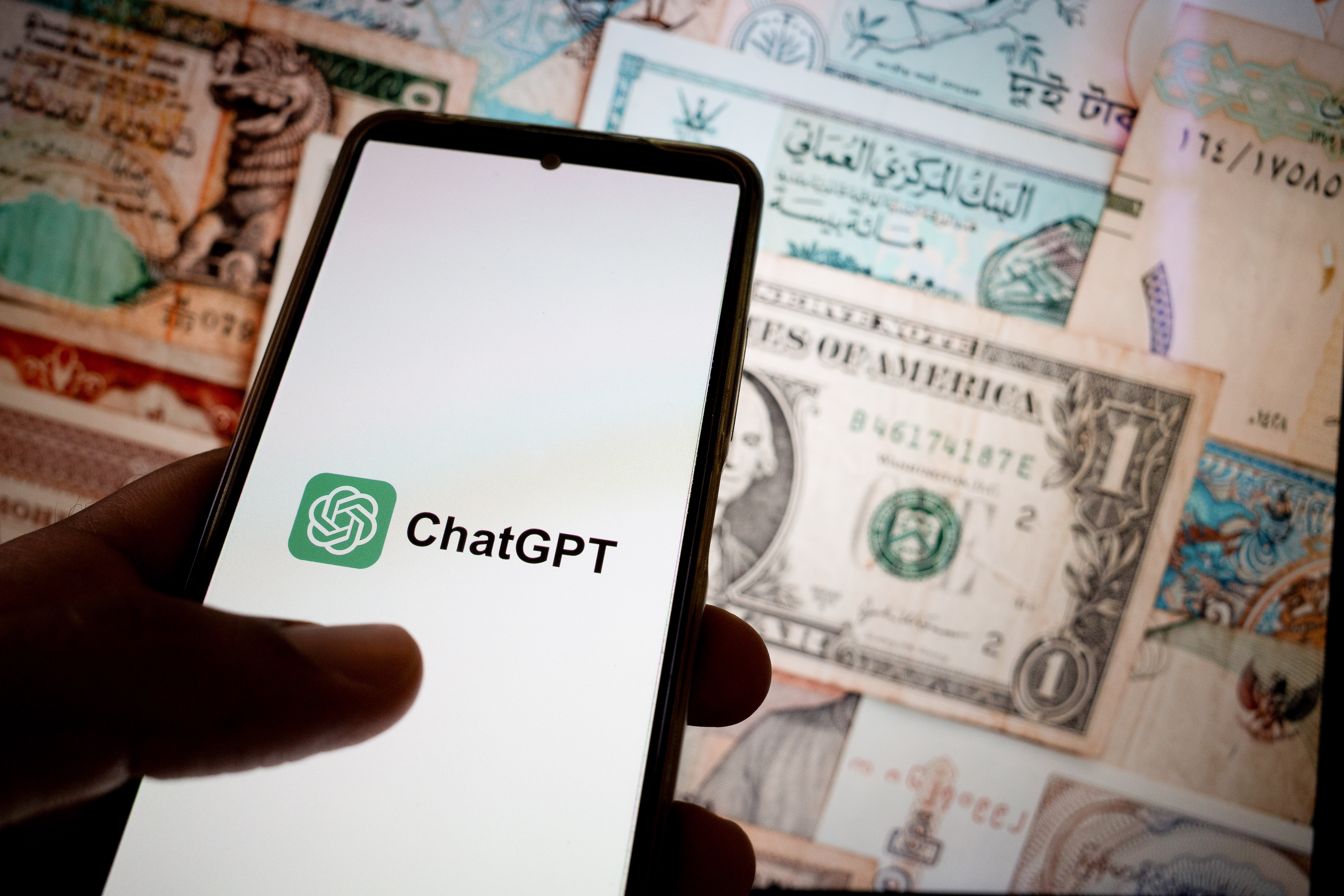 Phone displaying "ChatGPT" held in front of various international banknotes