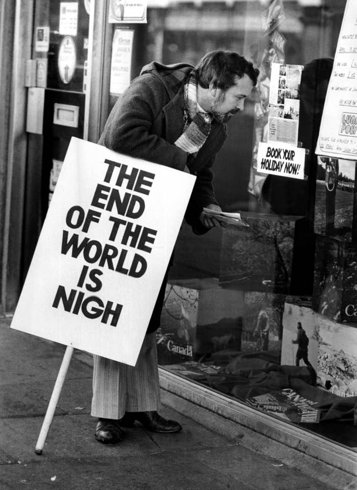 Person holding "The End of the World is Nigh" sign, peering into a travel agency window displaying holiday advertisements