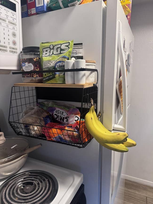 Kitchen scene with a black wire basket holding snacks, spices, bananas, and paper towels on the fridge. Stove with pot in foreground