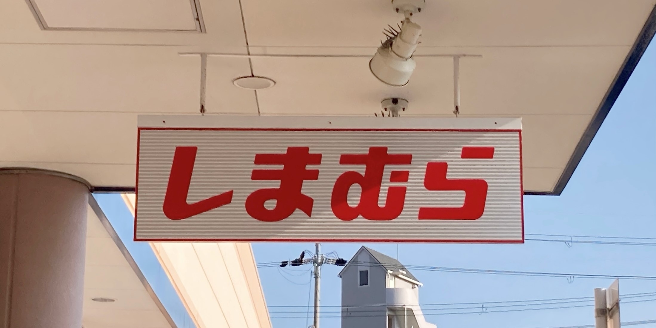 Store sign with Japanese characters, likely indicating "Shimamura." It's attached to an exterior ceiling with a partial view of a building and clear sky