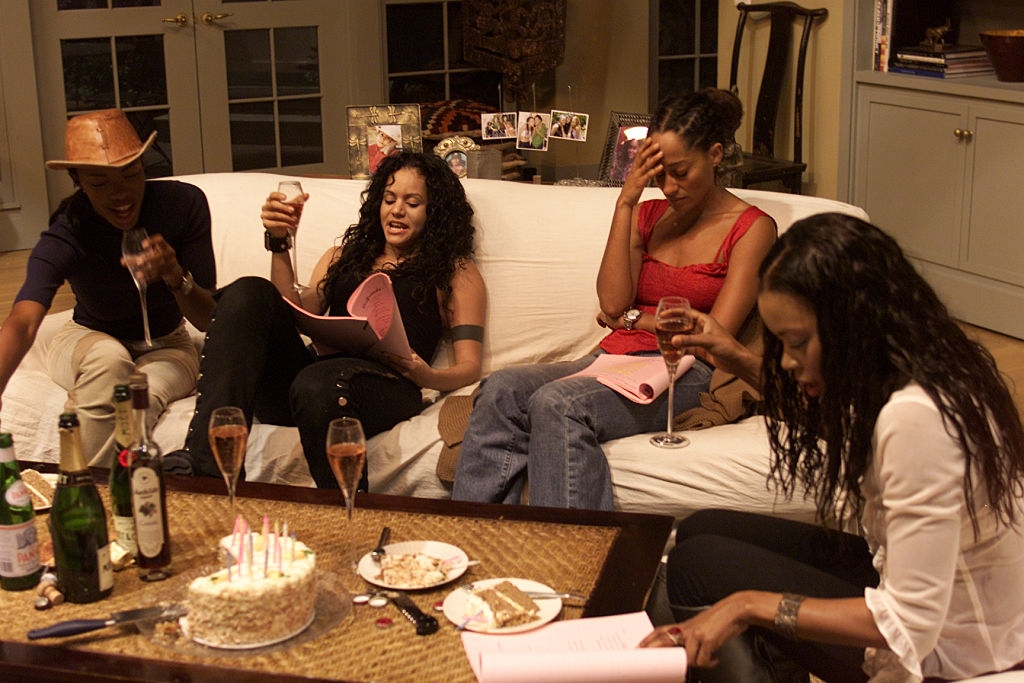 Four people gather around a table with a cake and drinks, reading scripts and laughing in a casual setting