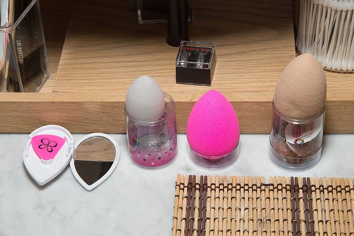Three makeup sponges in holders on a countertop, ranging from light grey to bright pink to tan, with a compact mirror open nearby