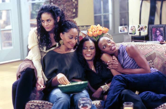 Four women sitting on a couch in a living room, smiling and interacting warmly, suggesting a scene from a TV show or movie