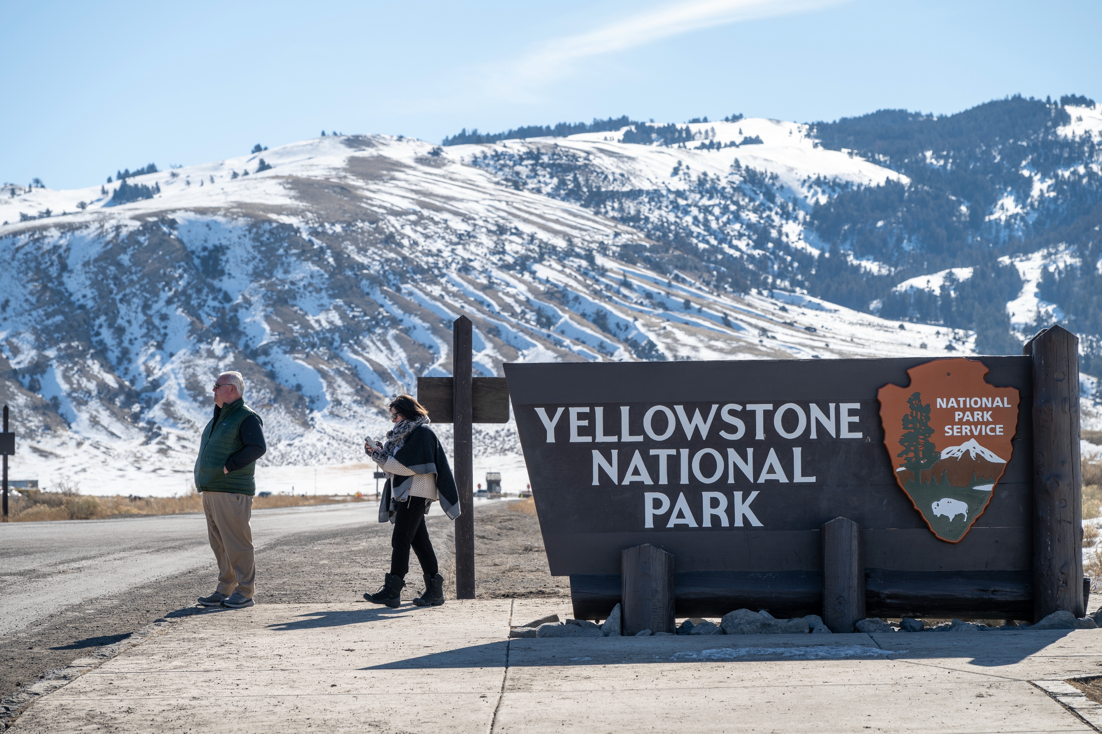 People standing near the Yellowstone National Park entrance sign with snowy mountains in the background