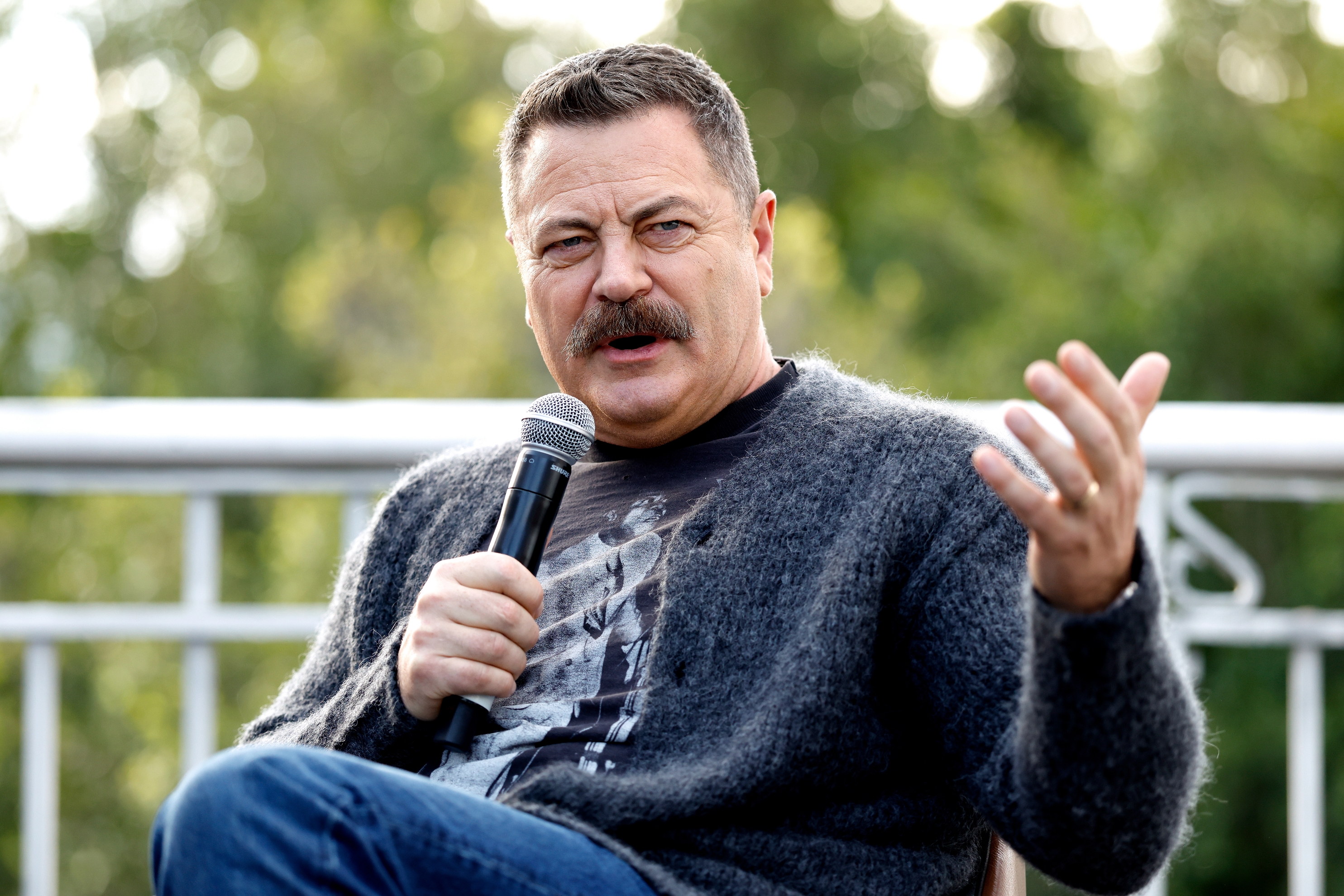 A person with a mustache speaks into a microphone while gesturing. They are wearing a casual outfit with a graphic T-shirt and a cardigan