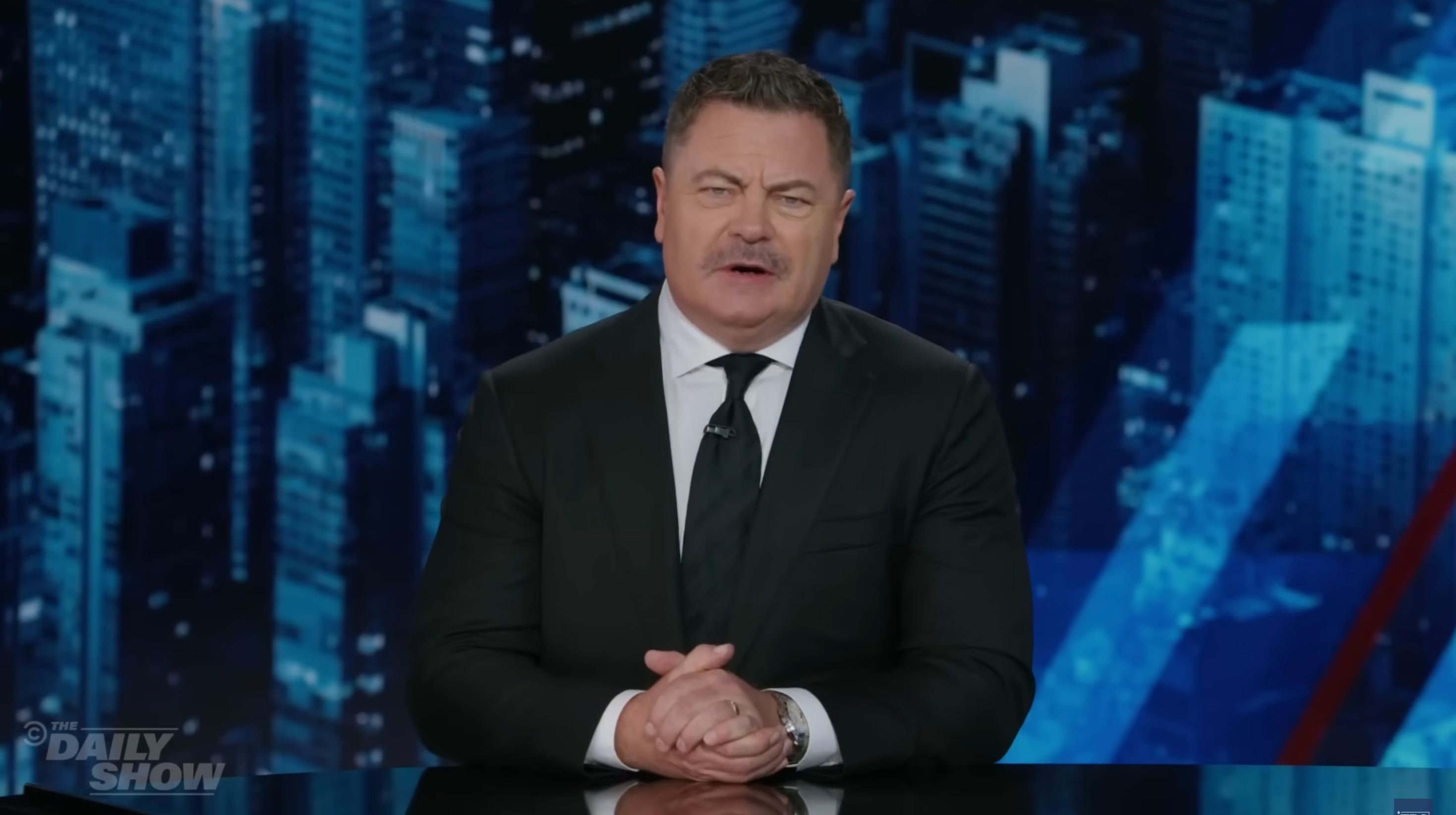 Person in suit speaking at a desk with a cityscape backdrop, possibly on a talk show or news segment