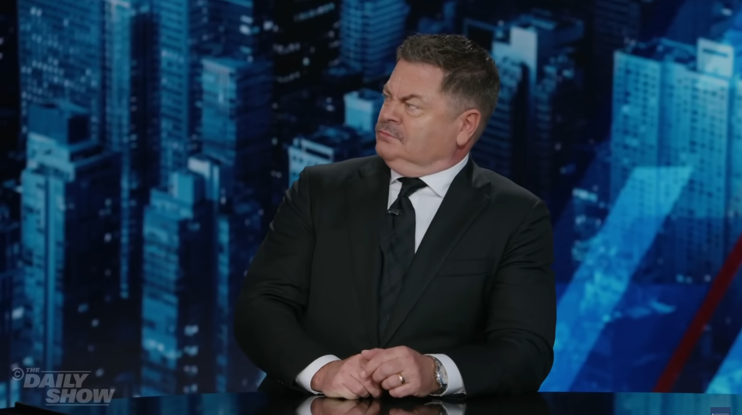 A person in a suit sits at a news desk, looking sideways with a serious expression, against a cityscape backdrop