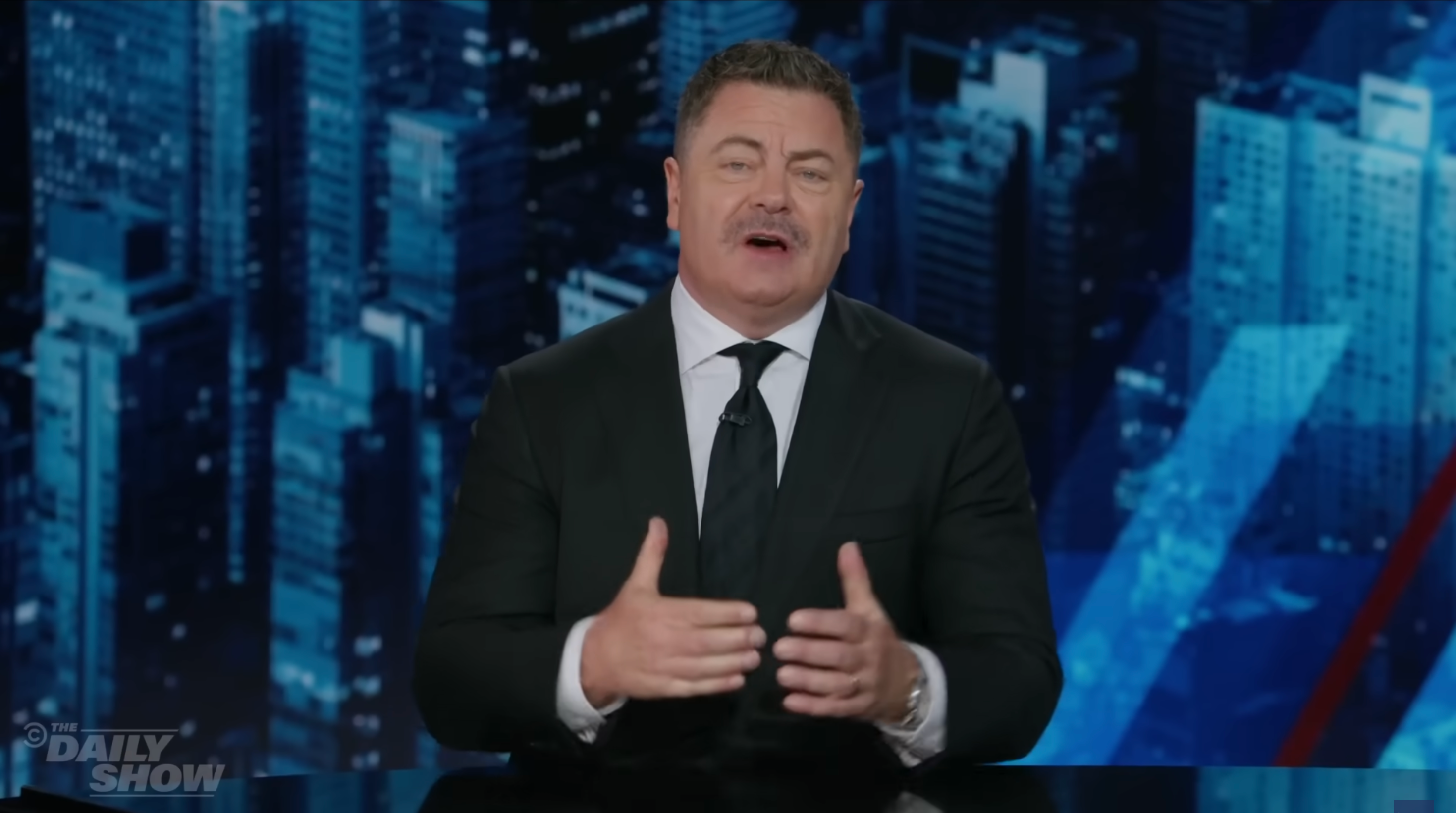 A man in a suit gestures while speaking at a news desk, with a cityscape in the background