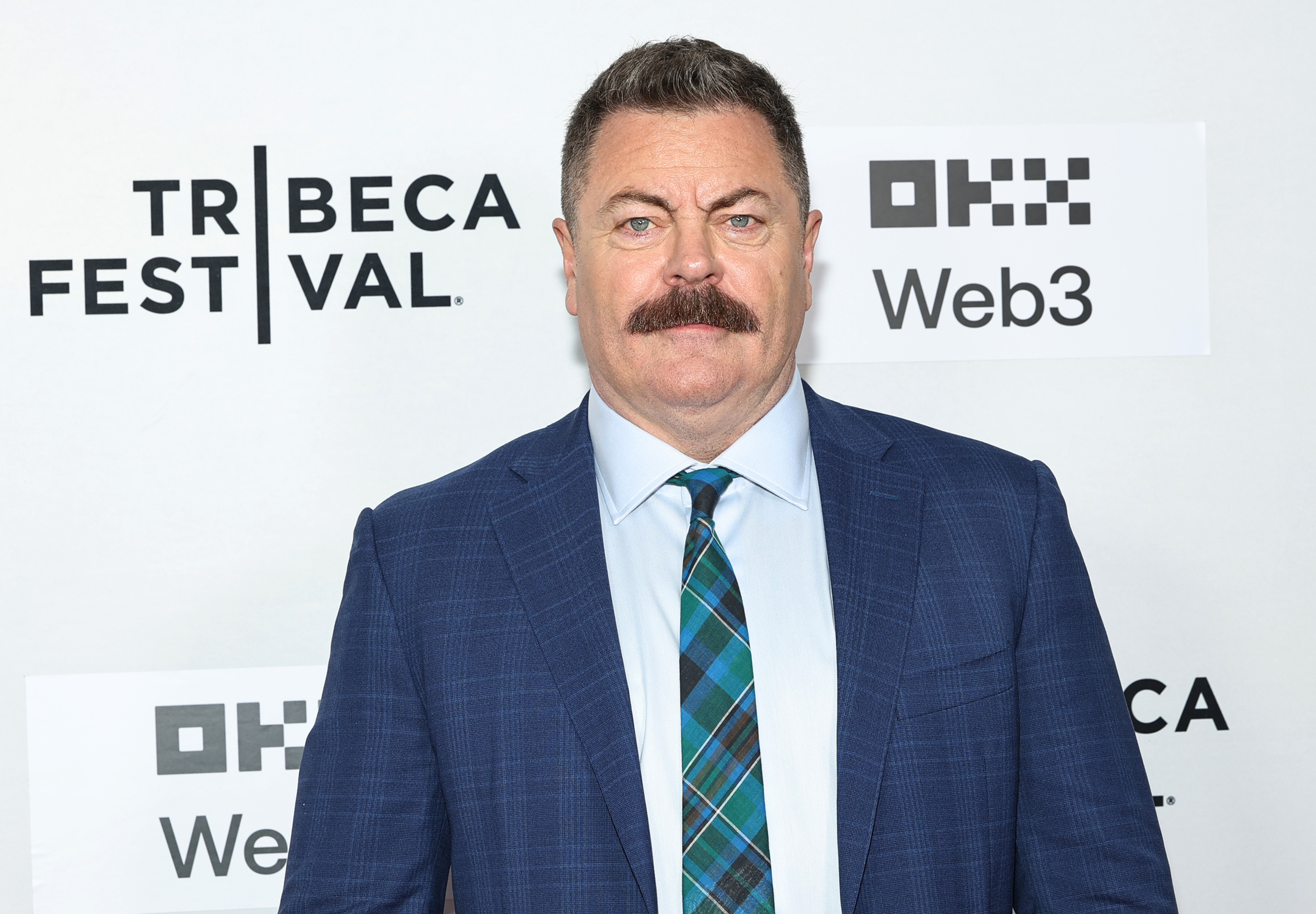 A person on a red carpet at the Tribeca Festival, wearing a blue plaid suit and tie, standing in front of a step-and-repeat banner