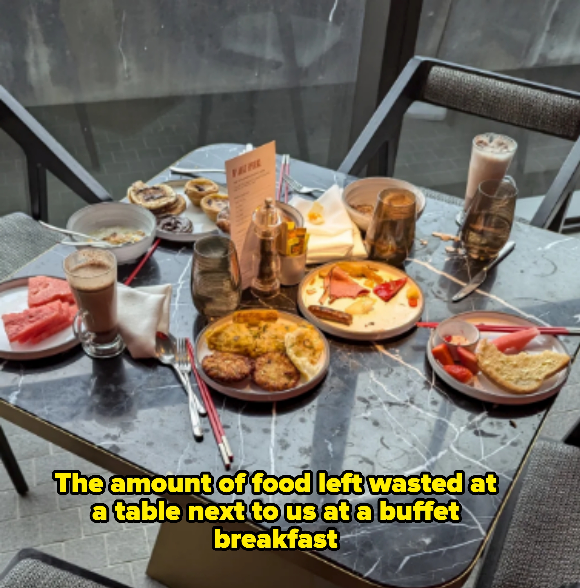 A restaurant table with a variety of breakfast foods, including watermelon slices, pastries, pancakes, yogurt, and two glasses of chocolate milk. Caption notes that a family at the buffet left all that food behind
