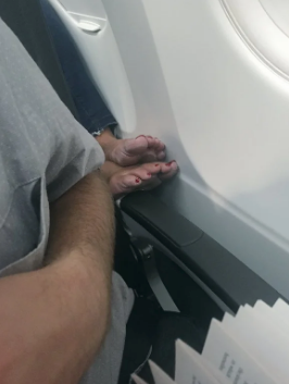 A person on an airplane resting their bare feet on the armrest of the person in front of them