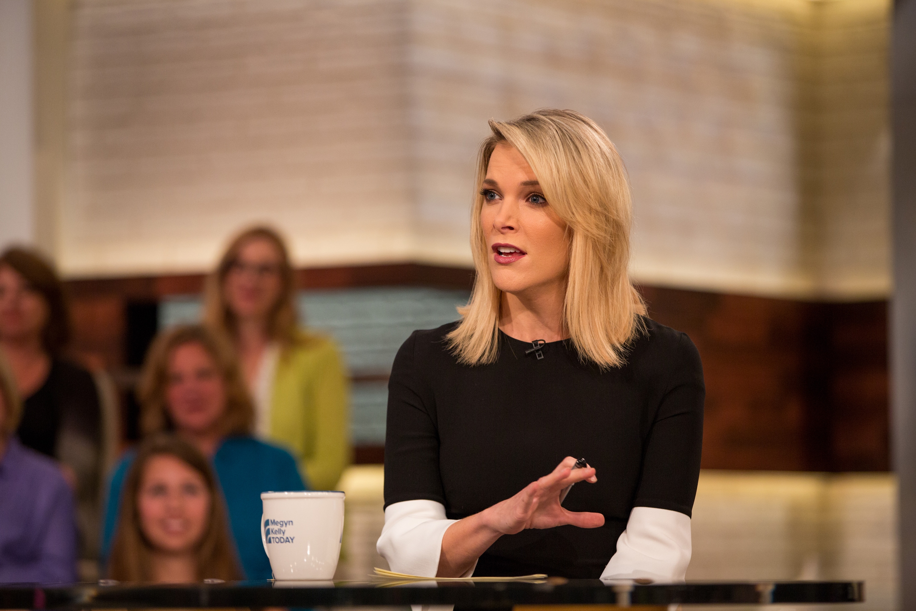 A person sitting during a talk show, gesturing with one hand, with a studio audience in the background