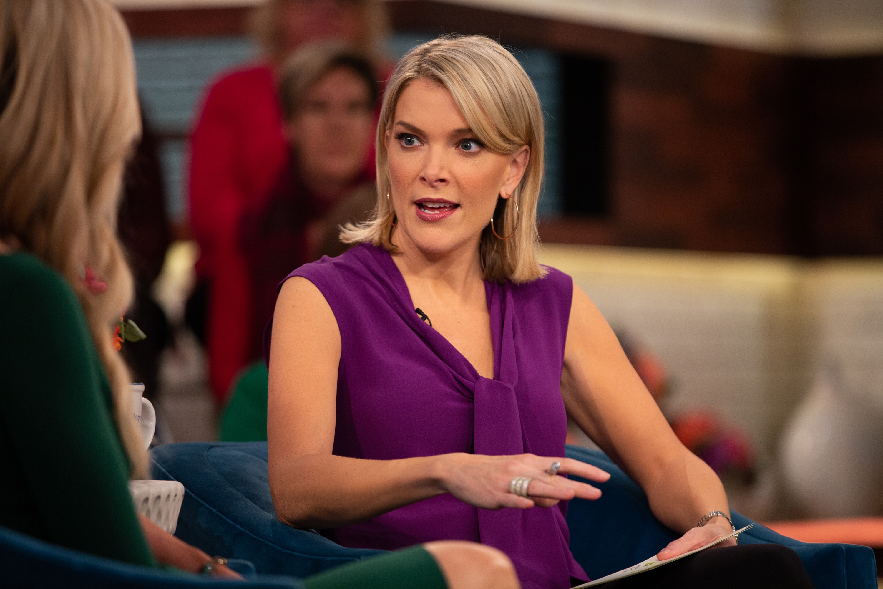 A person speaks animatedly on a talk show set, wearing a sleeveless top and seated in a brightly lit studio