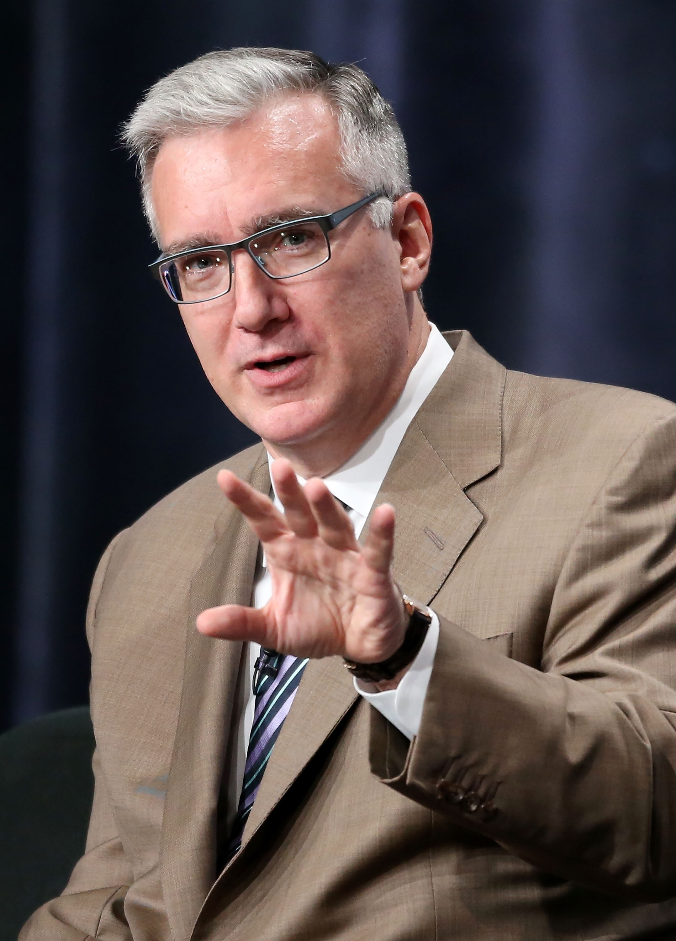 Person in a professional setting wearing a suit and tie, speaking with a hand gesture