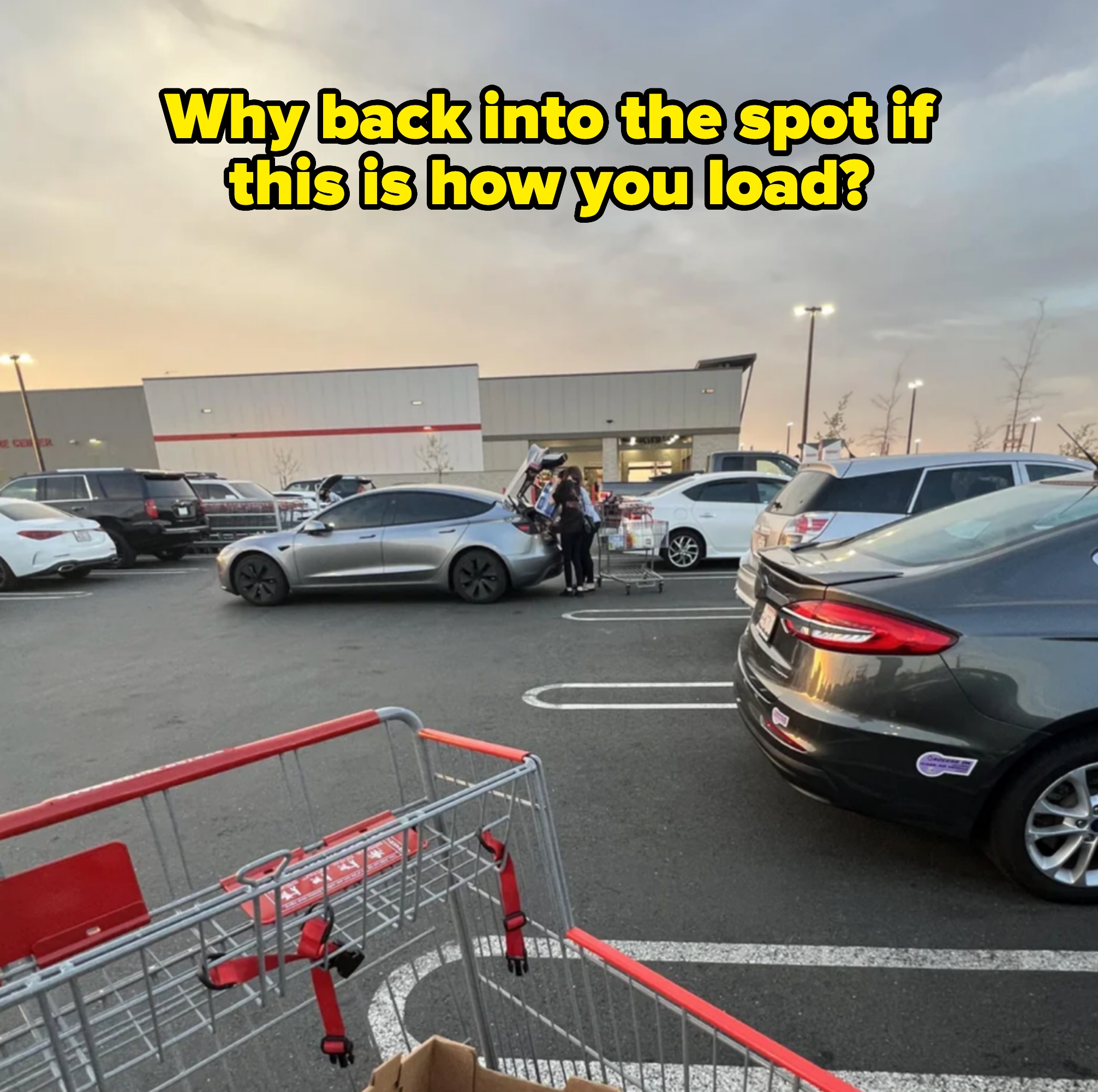 A person loads groceries into their car, which is parked in the middle of the driving path in the parking lot