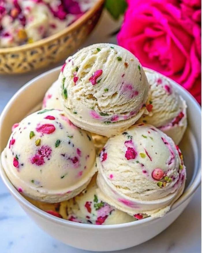 Bolas de helado en un tazón, decoradas con pétalos y trozos de frutas. Fondo con flores rosadas