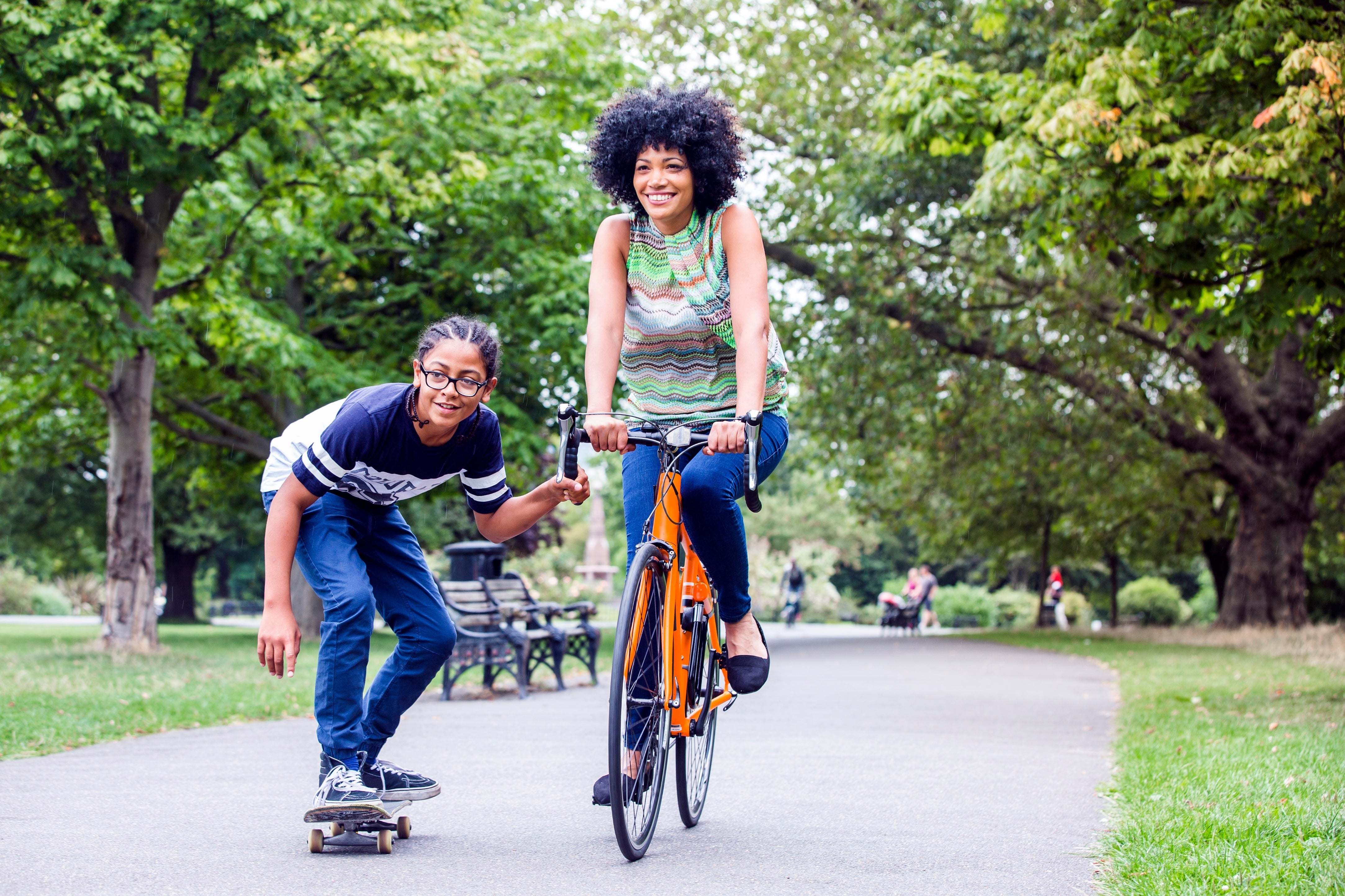 Kvinne som sykler og smiler, med en gutteskateboarding sammen med en parksti, omgitt av grøntområder