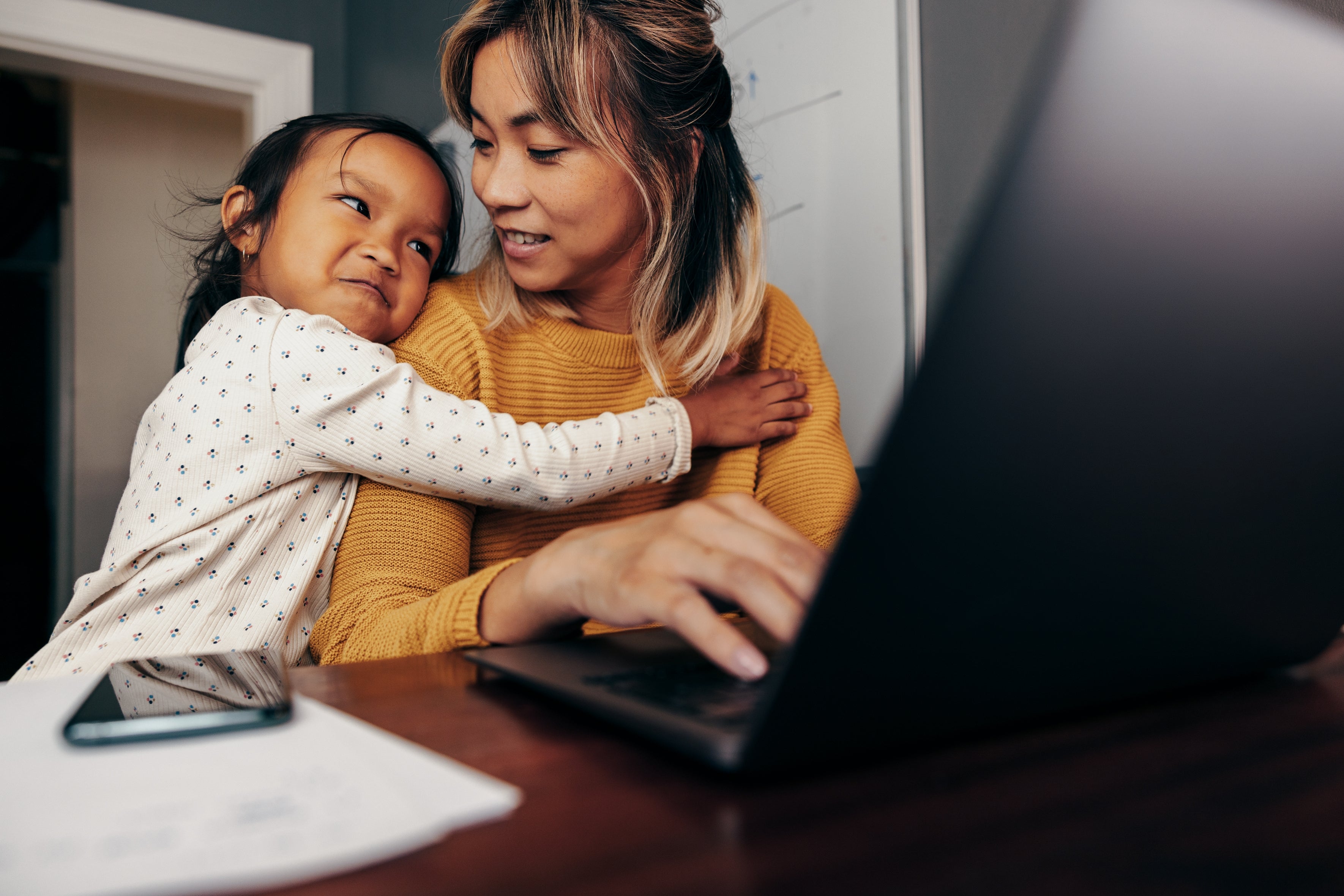 Barn som klemmer en smilende voksen som jobber på en bærbar datamaskin på et skrivebord med papirer og en telefon. Koselig hjemmeinnstilling
