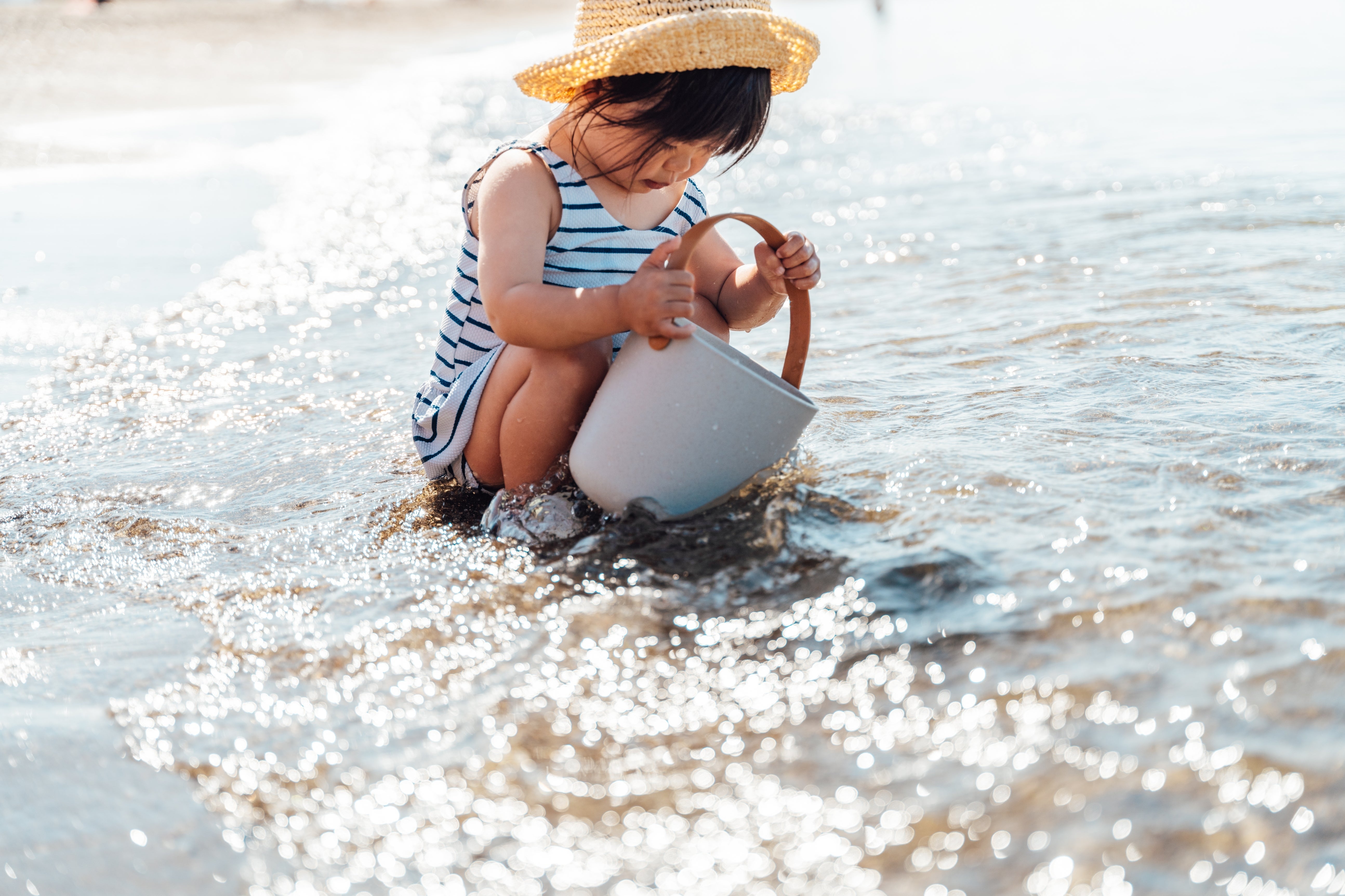 Barn på stranden som leker med bøtte på grunt vann, iført stripete antrekk og stråhatt