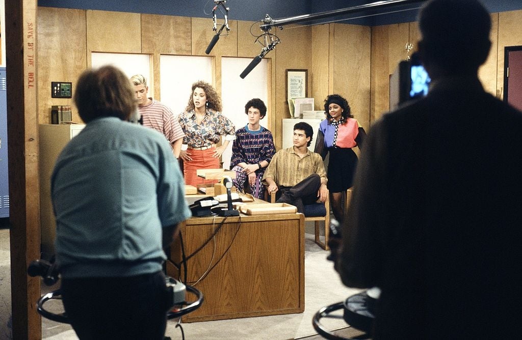 People filming a TV country acceptable successful an office, featuring actors successful 1980s attire and unit members operating cameras