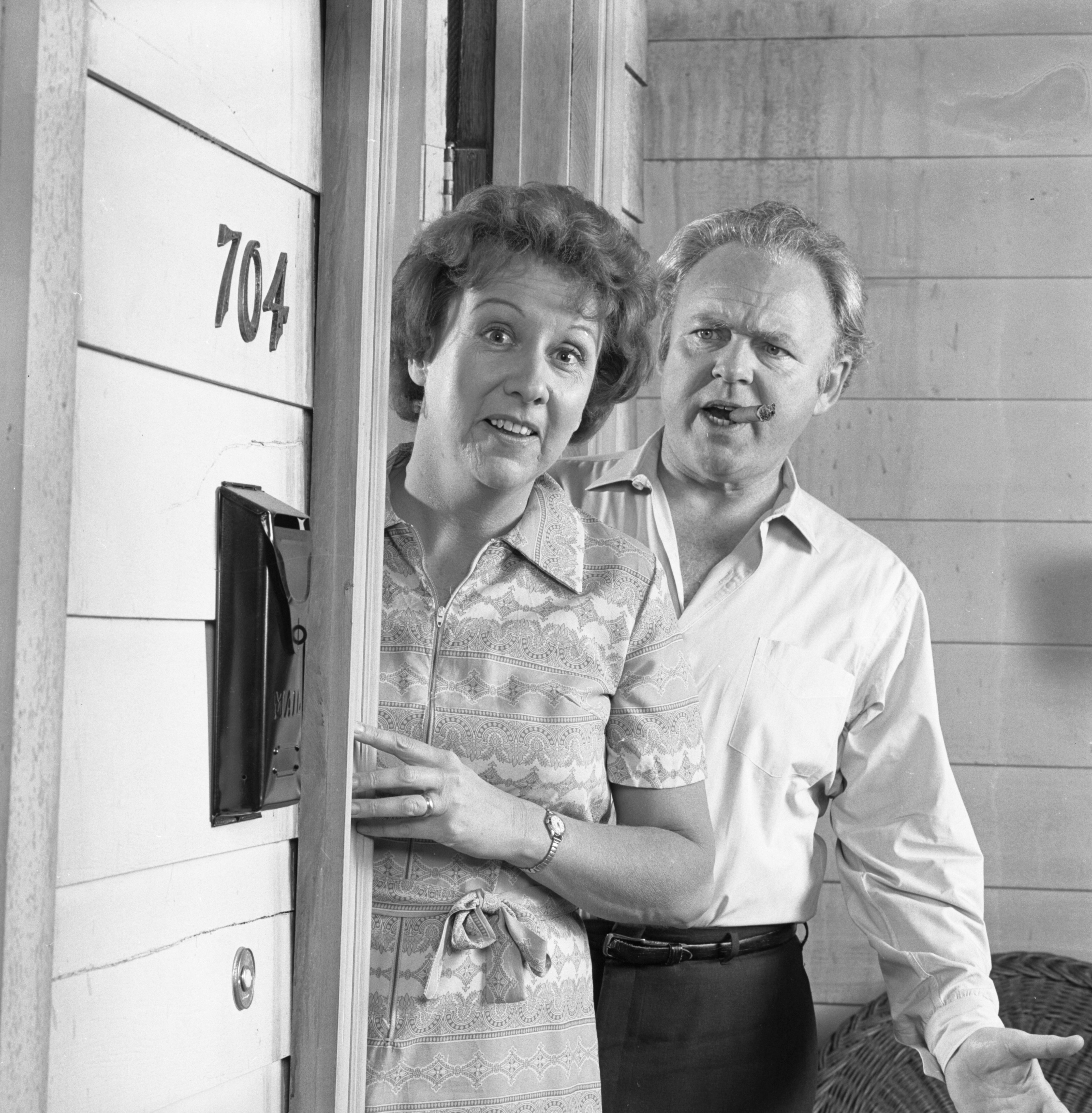A mates stands successful a doorway labeled "704," looking surprised; the pistillate wears a patterned dress, and the antheral is successful a collared shirt