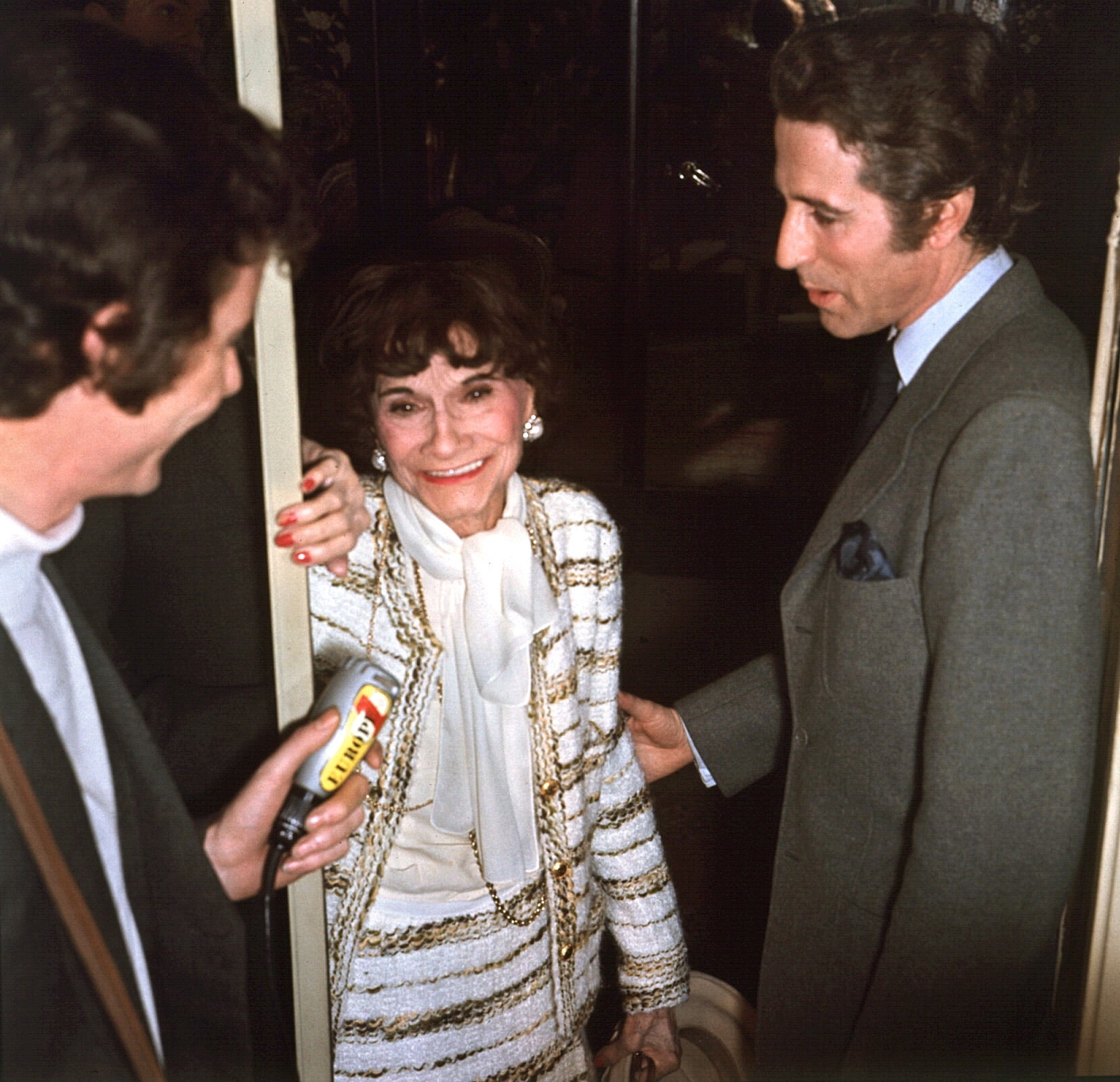 A pistillate successful an elegant, patterned suit smiles portion speaking with 2 men, 1 holding a microphone, astatine an event