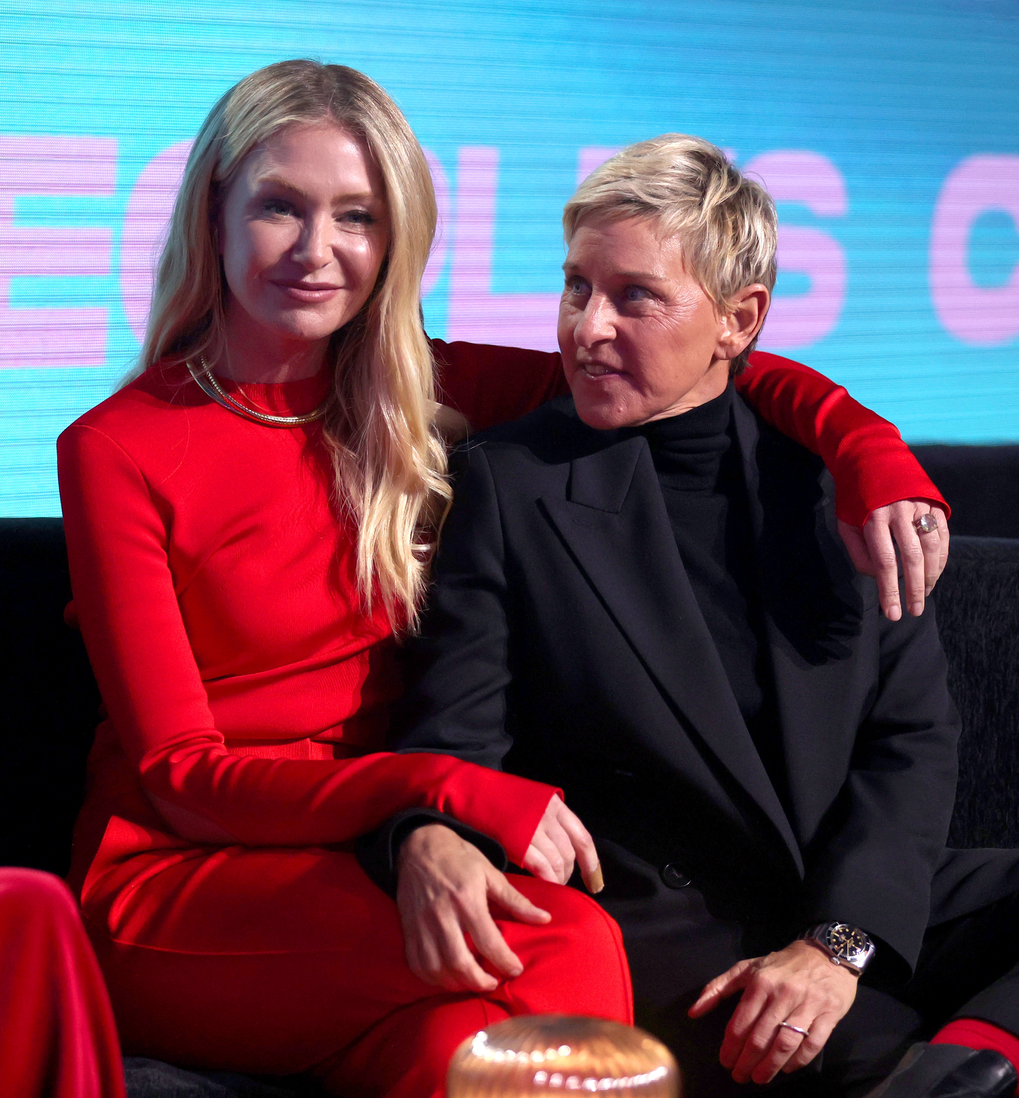 Two people sitting together on a stage. One wears a red outfit, and the other wears a black suit. They appear relaxed and engaged in conversation