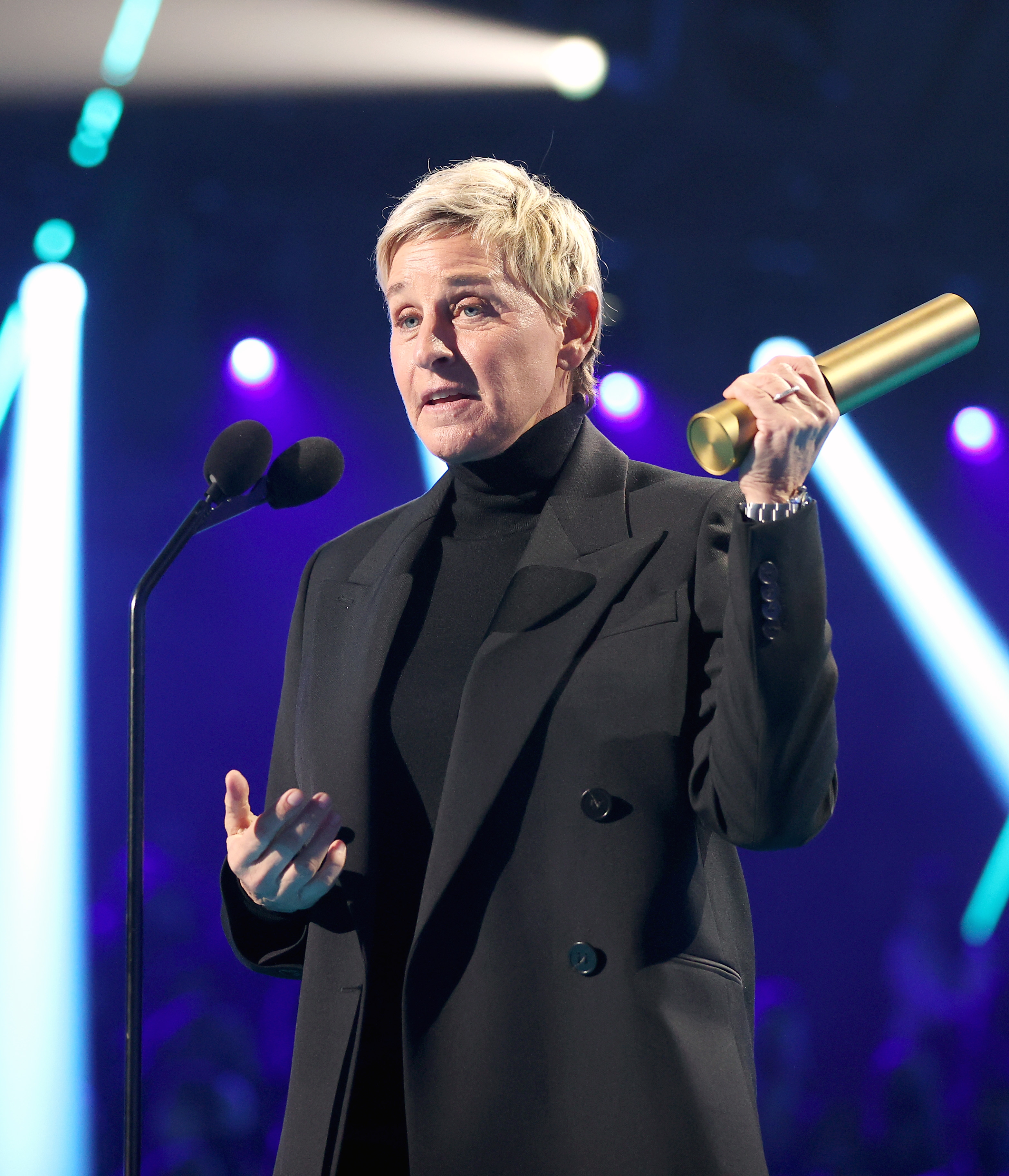 Person in a suit holds a cylindrical award, speaking into a microphone on stage with bright lights in the background