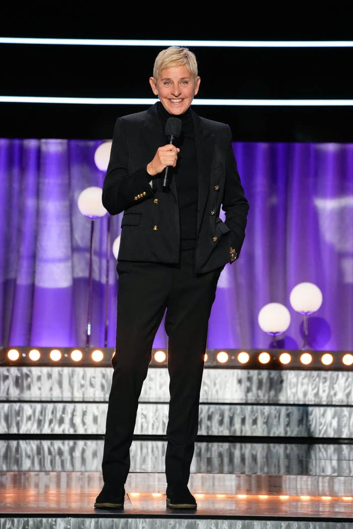 Person in a sleek black suit and turtleneck holds a microphone on stage with circular lights and a curtain background