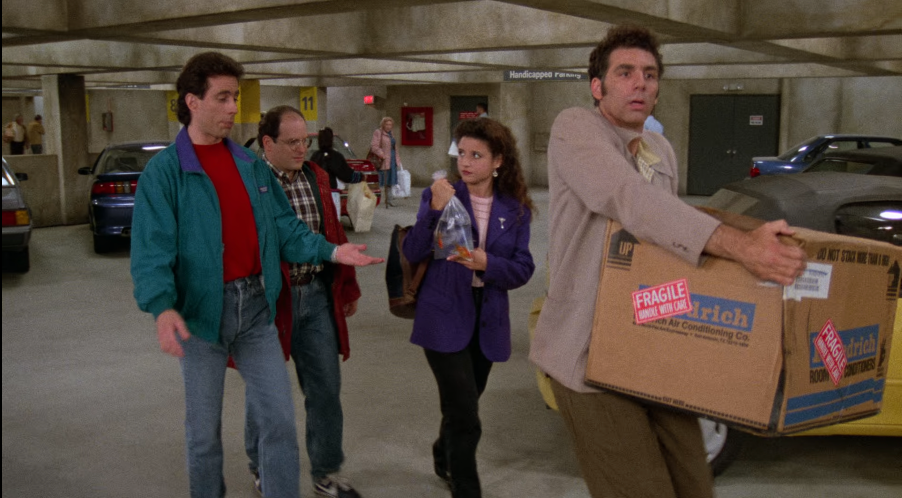 Jerry, George, Elaine, and Kramer walk through a parking garage. Kramer carries a large box marked &quot;fragile,&quot; and Elaine carries a goldfish in a plastic bag