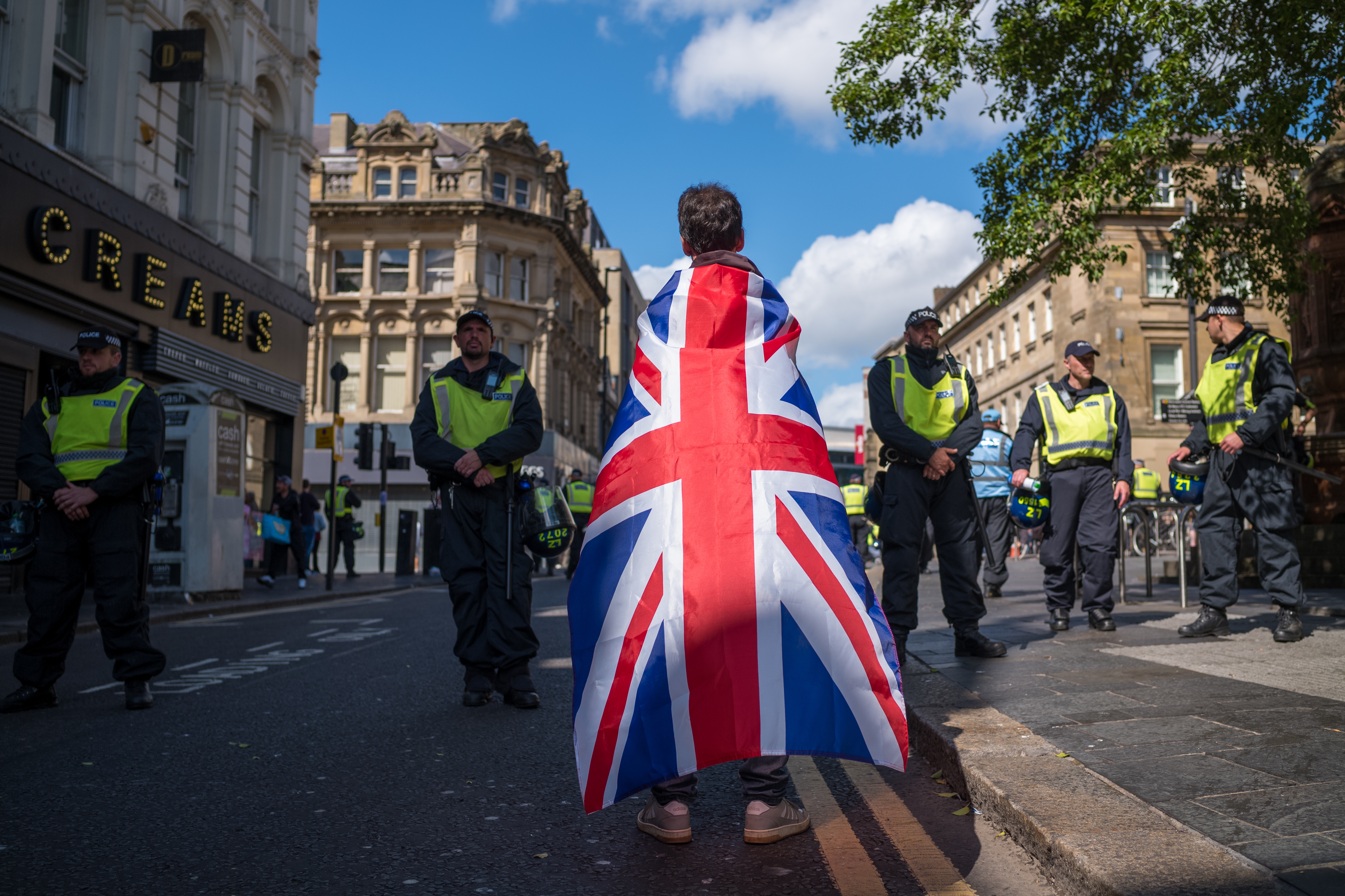 Person draped in Union Jack facing police officers in a city street, during a public gathering