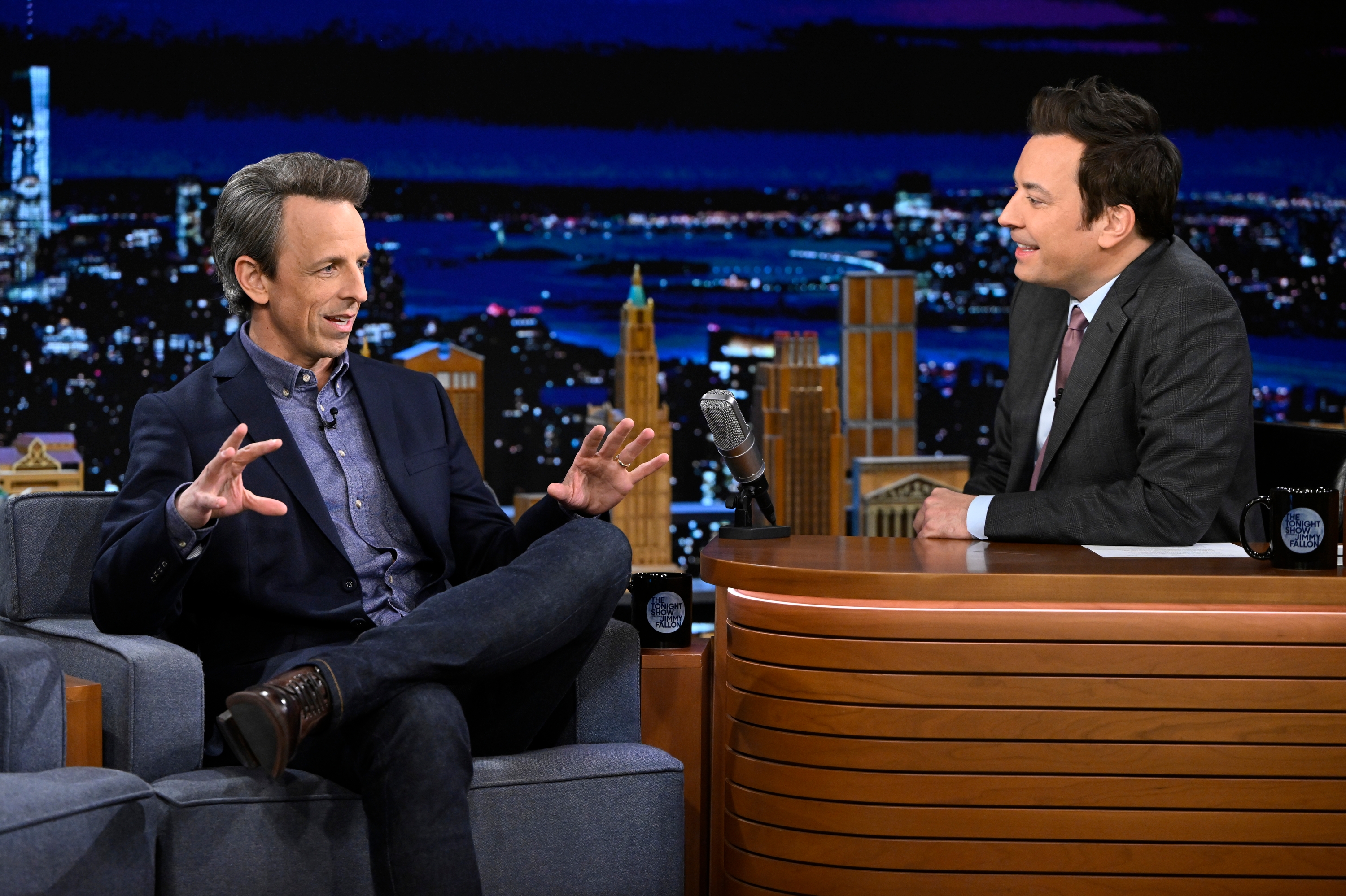 Two talk show hosts sit and converse on a late-night set, one gesturing animatedly, the other listening attentively behind a desk with a microphone
