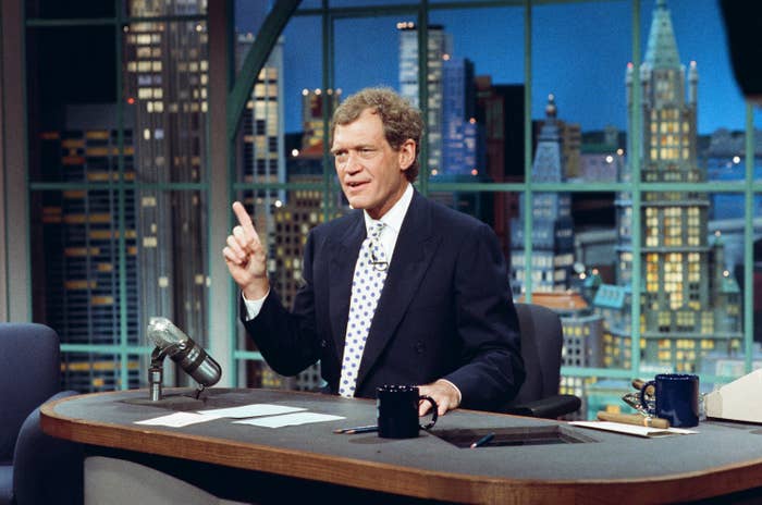 Talk show host gestures while sitting at a desk with papers and a microphone, set against a city skyline backdrop