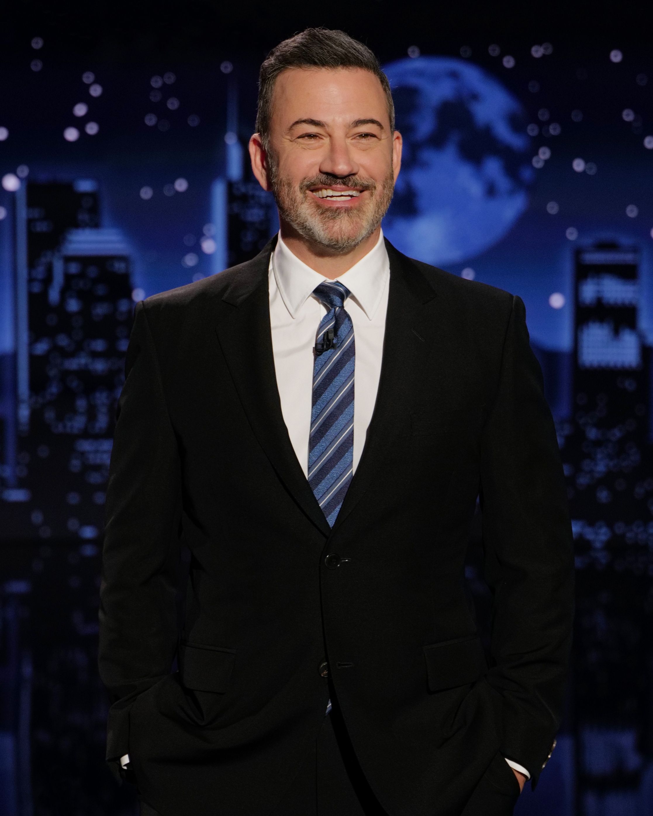 A person in a suit and tie stands on a stage with a cityscape backdrop and a large moon, smiling with hands in pockets