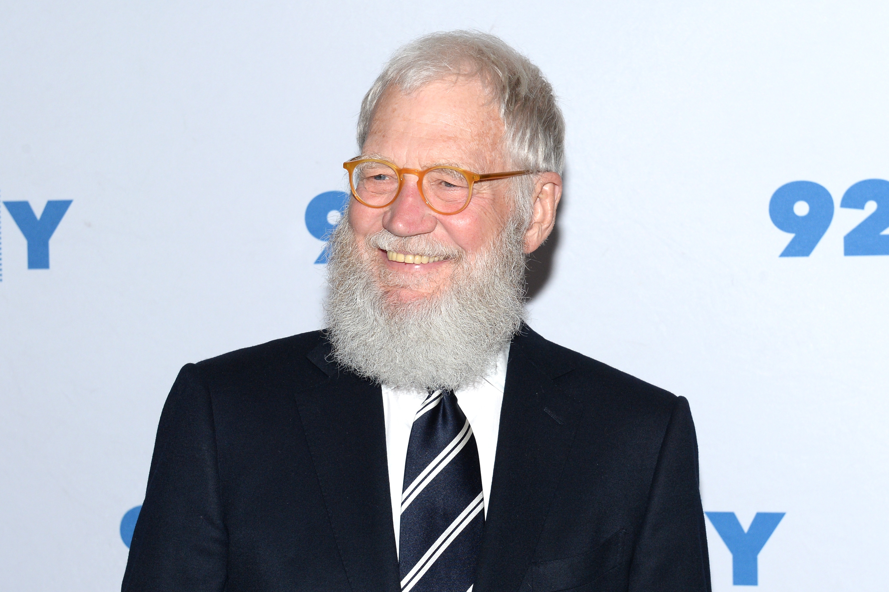 Person with a white beard wearing glasses and a suit with a striped tie, smiling at an event against a plain backdrop
