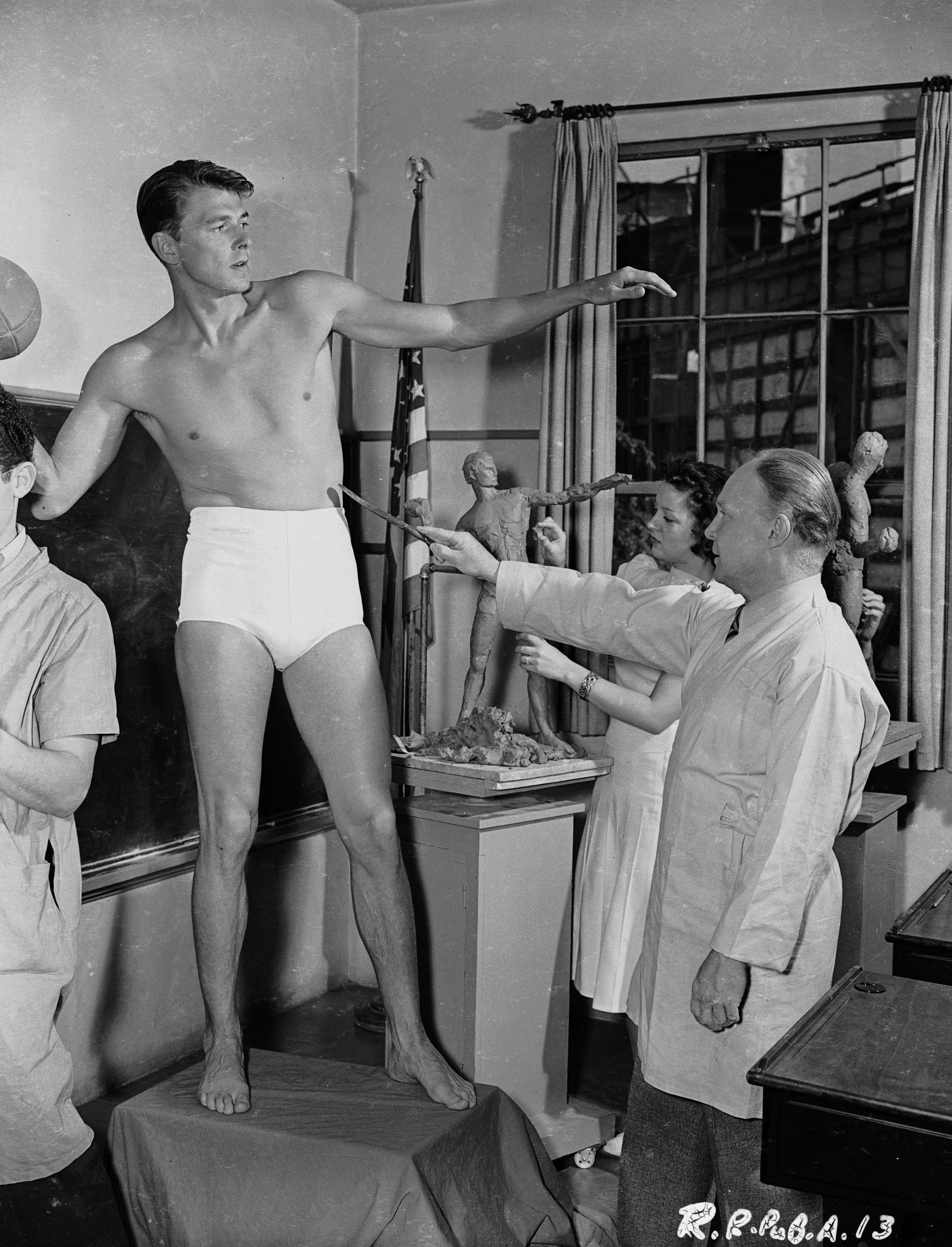 A antheral poses arsenic an artist's exemplary successful a classroom, surrounded by sculptors astatine work, with a teacher giving instructions