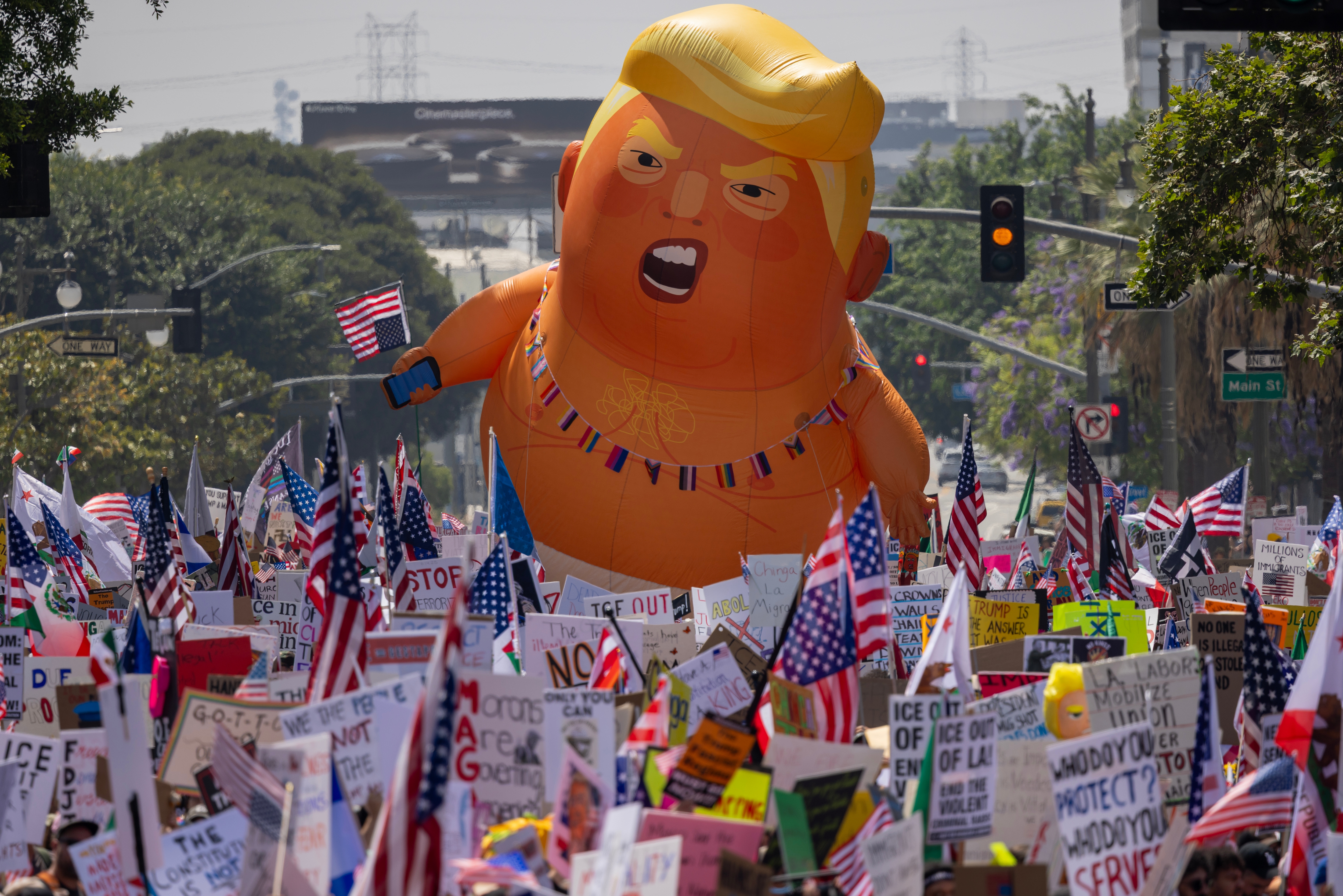 Protest with many people holding signs and flags, featuring a large inflatable balloon resembling a public figure in the background