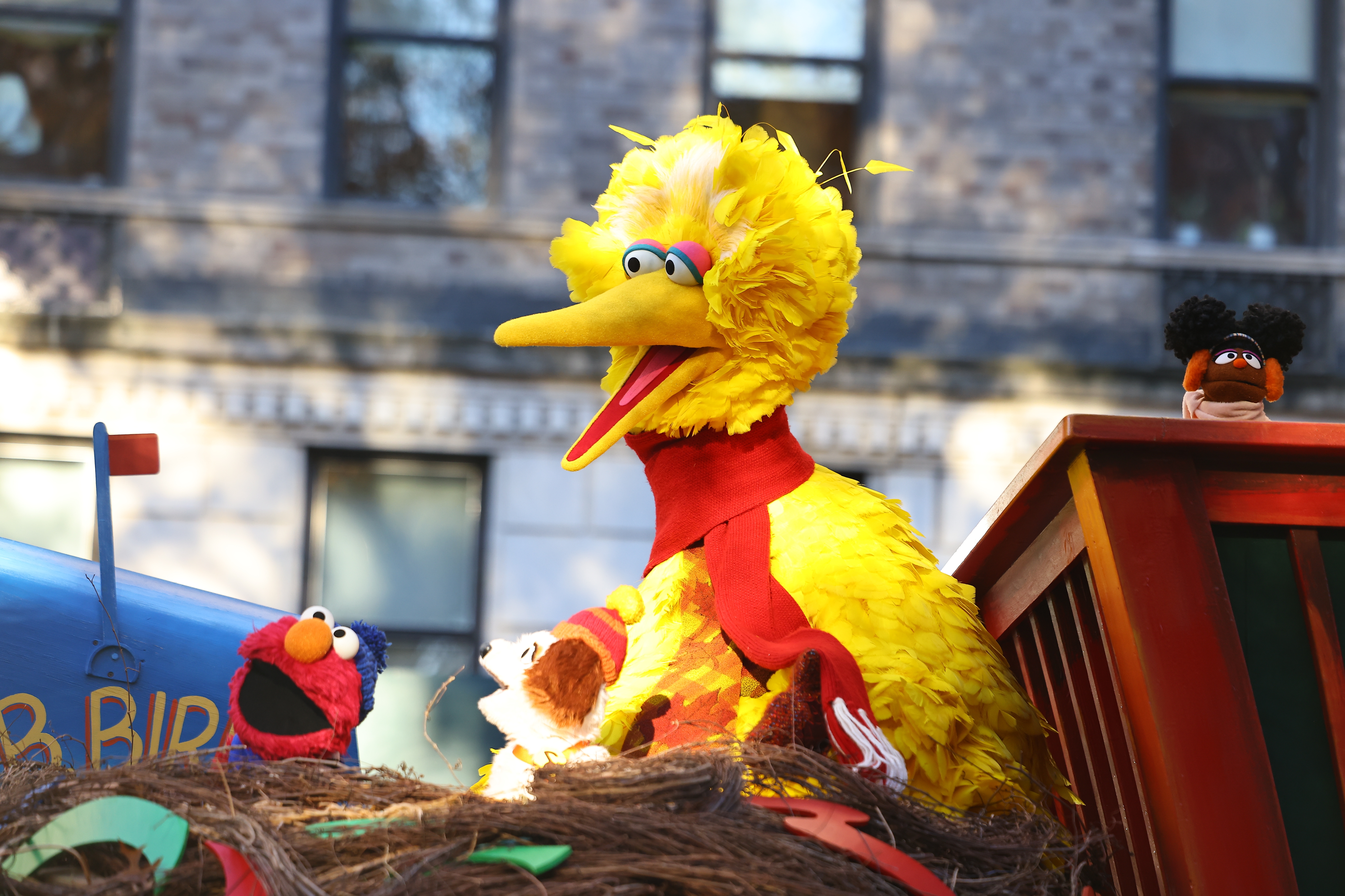 Big Bird and Elmo waving from a parade float decorated like a nest, in front of a building